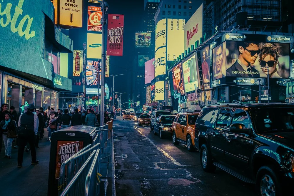 Nighttime city street scene with cars parked along the curb, bright digital billboards, and a crowd of people walking on the sidewalk.
