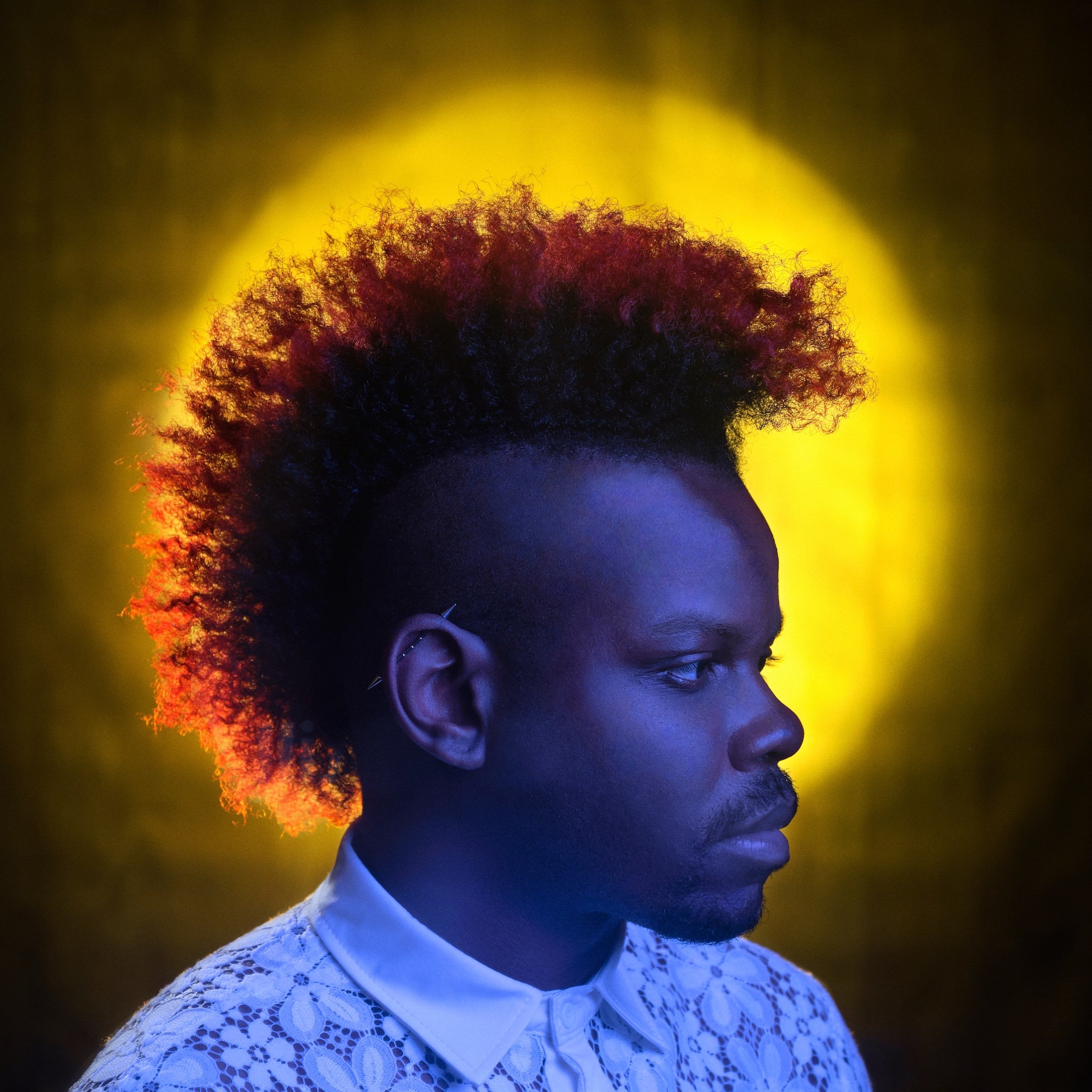 Side profile of a person with curly hair, wearing a white lace top, against a background with yellow and dark lighting.