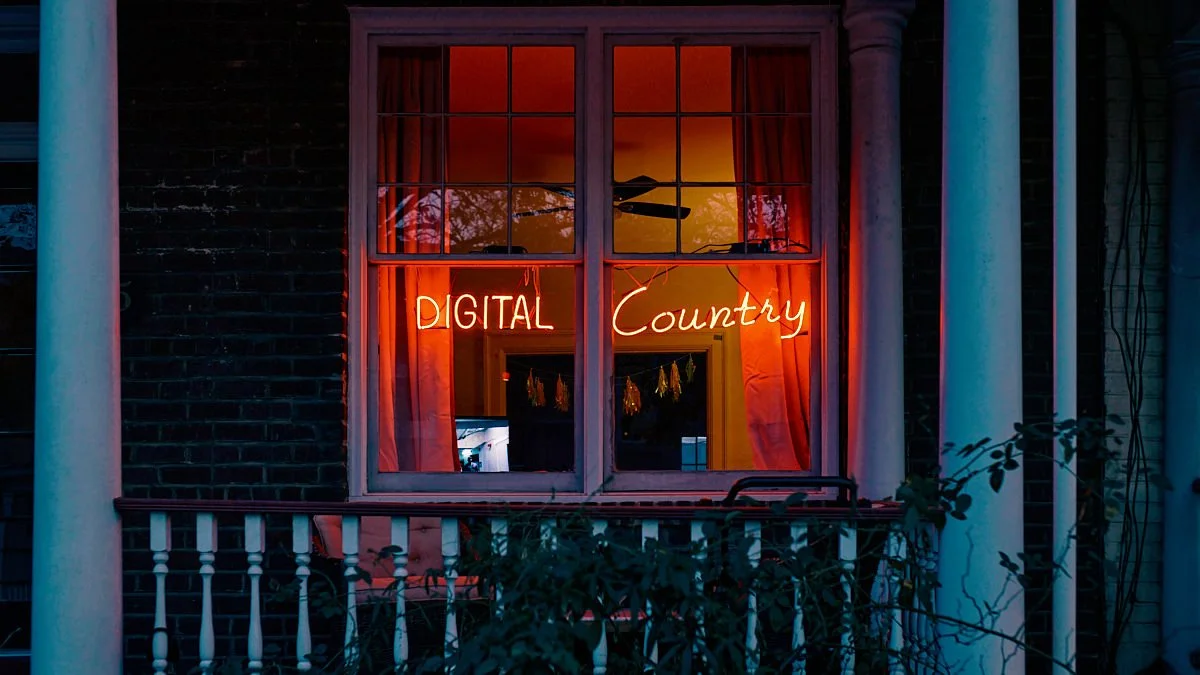 A window with neon signs reading 'DIGITAL Country' inside a dark house, framed by white columns and a porch railing with some greenery in front.