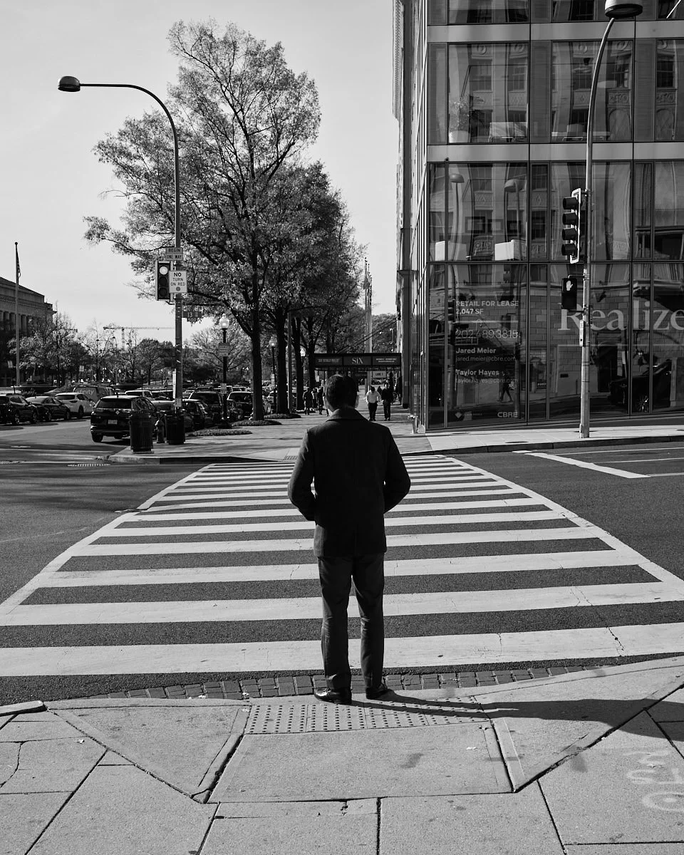 A man in a suit stands at a crosswalk on a city street, with trees, cars, and modern buildings in the background, visible in black and white.