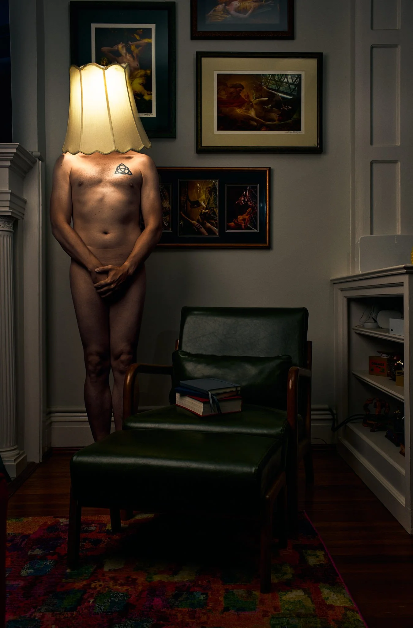 A surreal photo of a nude man standing in a living room with a lampshade covering his head. The man has a tattoo on his chest. The room has framed artwork on the wall, a chair with a book on it, and a colorful carpet on the floor.