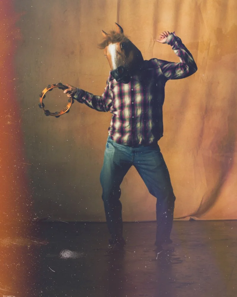 A person wearing jeans and a plaid shirt with the head of a horse, holding a tambourine in one hand and making a fist with the other, standing in front of a beige backdrop.
