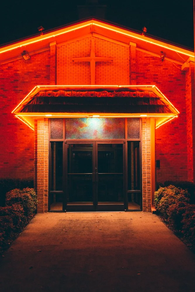 Night view of a church entrance with brick facade, illuminated by orange neon lights outlining the roof and awning, and a cross on the wall above the door.