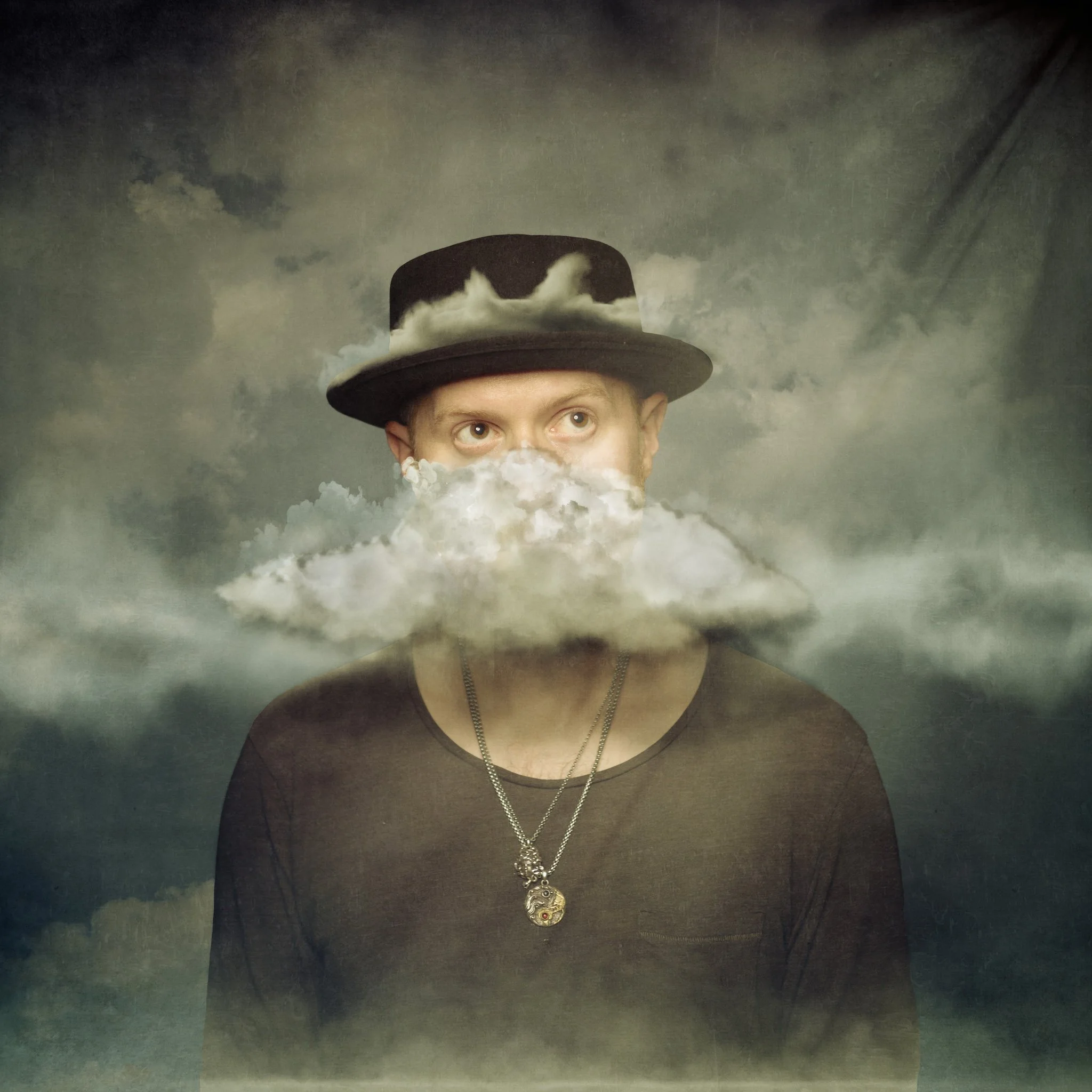 A person wearing a black top, a silver chain necklace, and a black fedora hat with clouds and sky reflected on the hat's surface.
