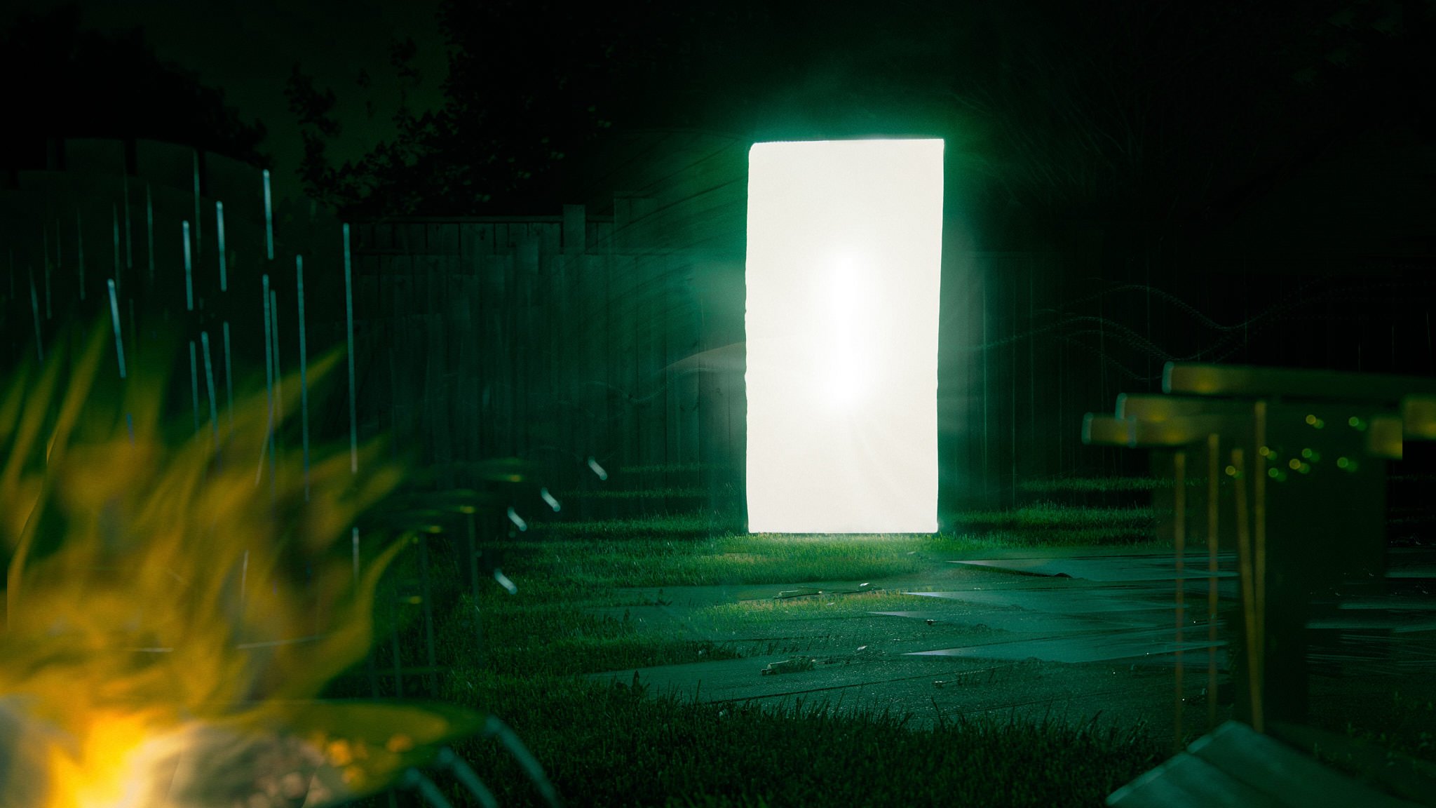 Brightly lit white rectangular screen or sign in a dark outdoor setting, with some out-of-focus grass and trees around, and a yellow object in the foreground.