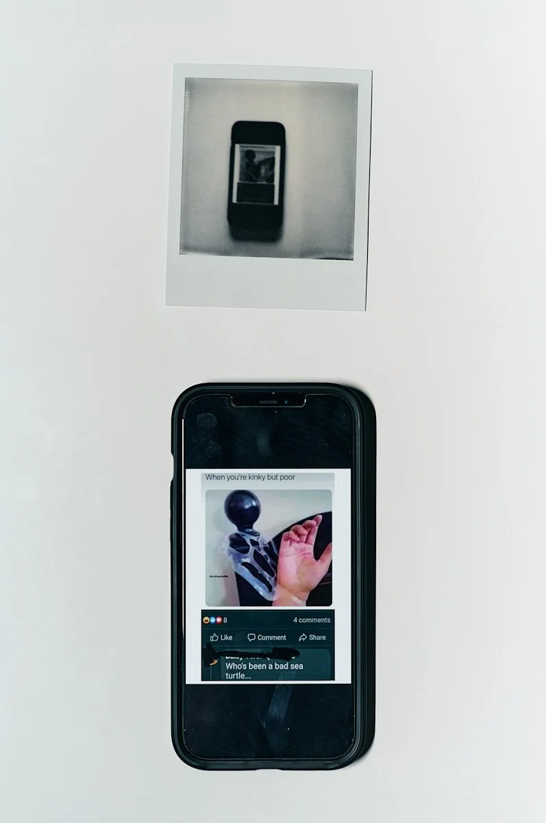 A white background with two object, one above the other. The object on top is a polaroid print of a cellphone displaying the same image as the object below it. On the phone screen is a social media post with likes and comments.