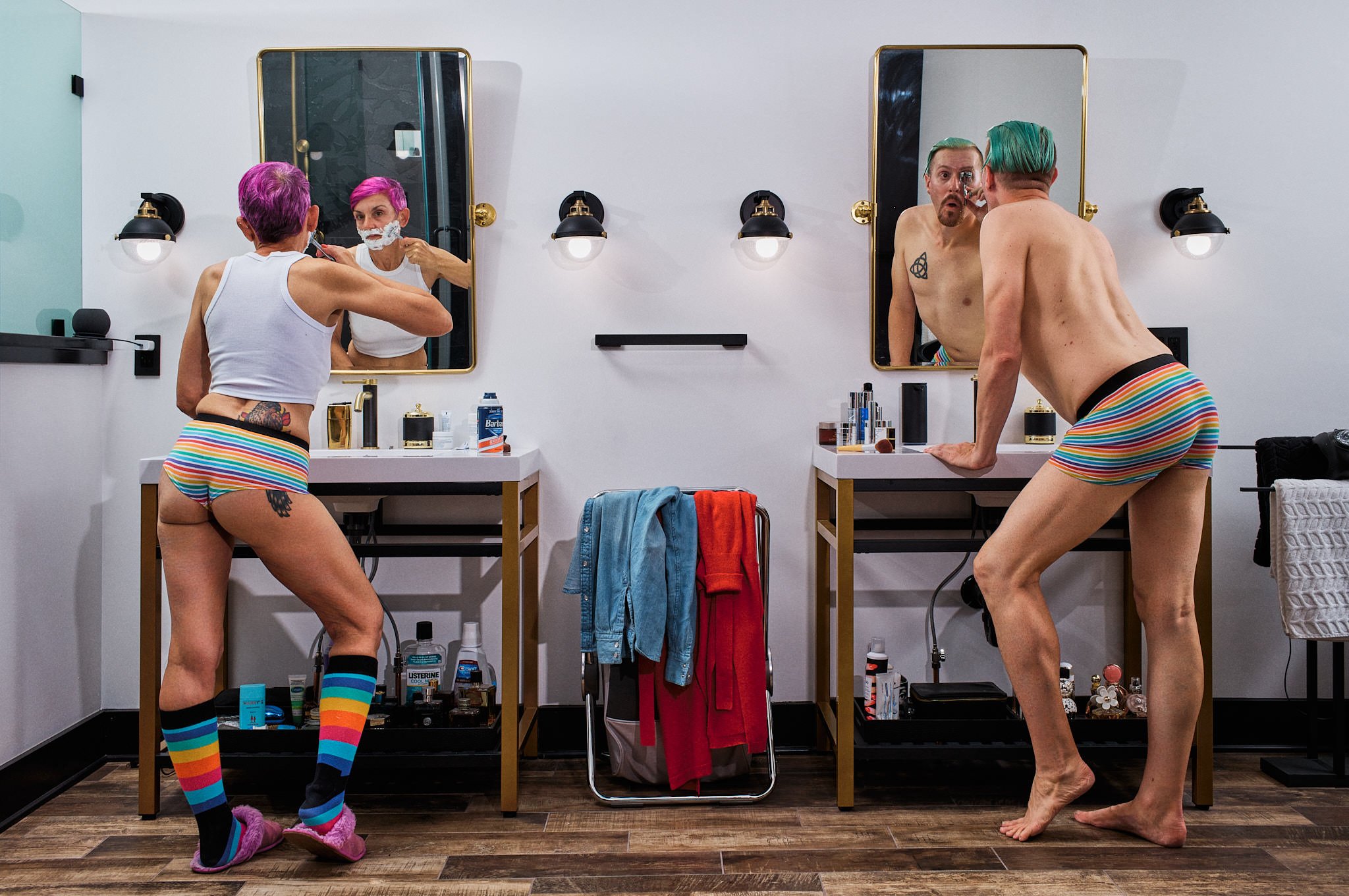 A man and woman with colorful hair and outfits, getting ready in a bathroom with mirrors and grooming supplies, the woman shaving and the man looking into the mirror.