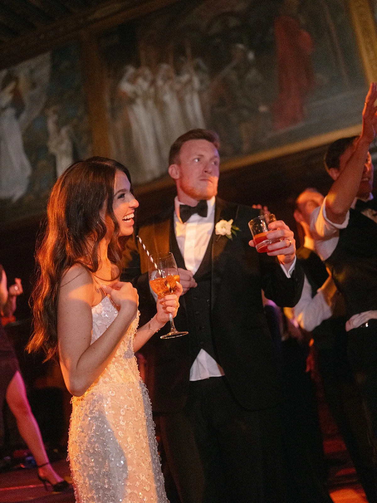 A bride and groom dancing and celebrating at their wedding reception, with the bride in a white dress holding a wine glass and the groom in a tuxedo holding a drink, surrounded by guests at the BPL.