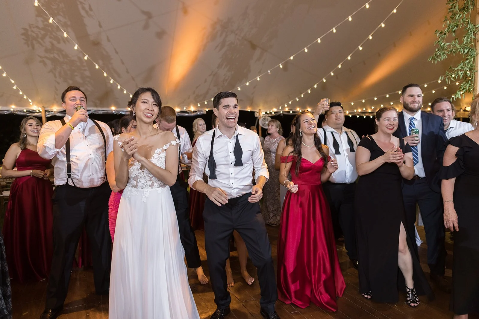 A group of people dancing and celebrating at a wedding reception under string lights, with the bride and groom in the center.