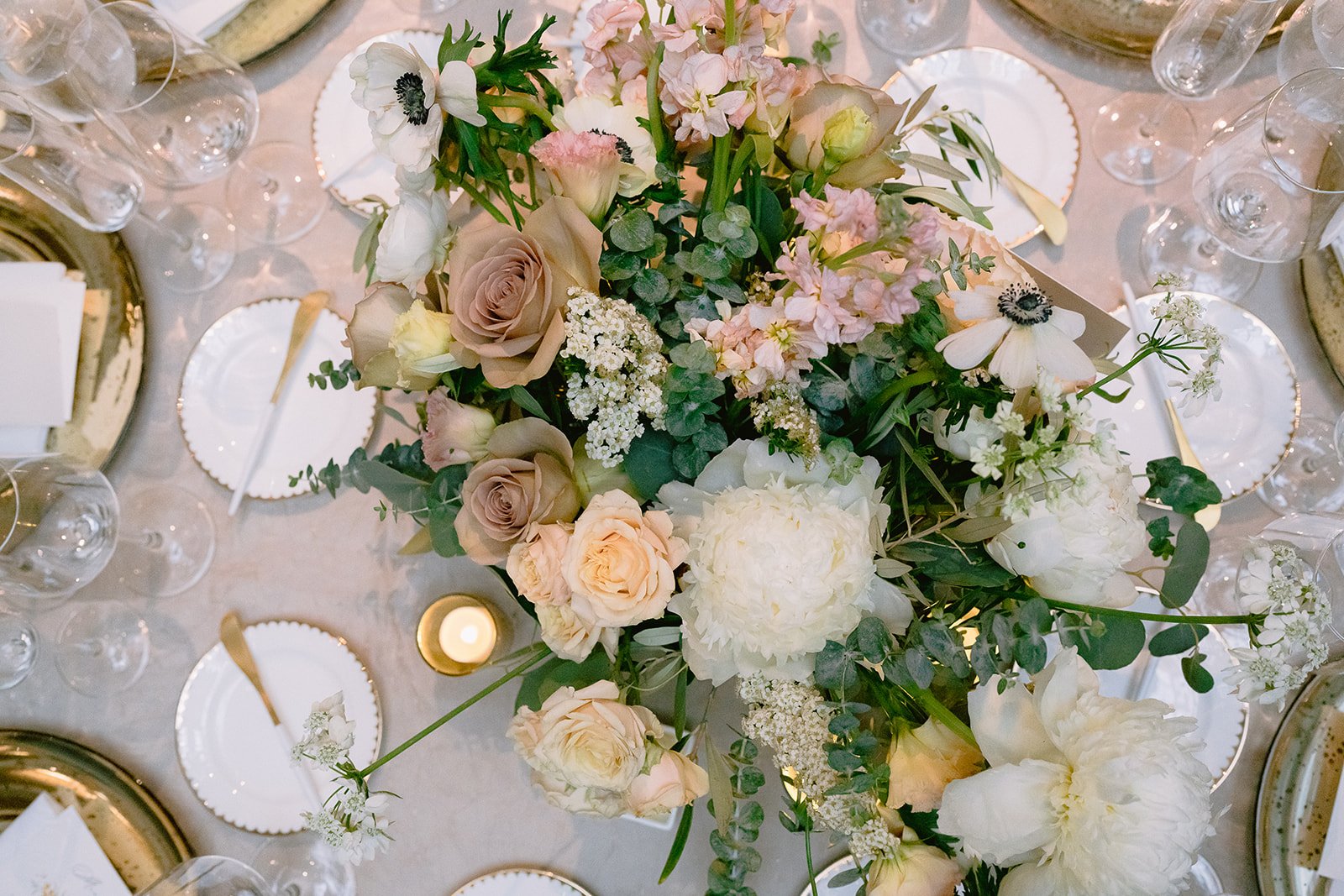 Elegant table setting with a floral centerpiece of cream, blush, and white flowers, surrounded by gold-rimmed plates, gold utensils, wine glasses, and a small candle.