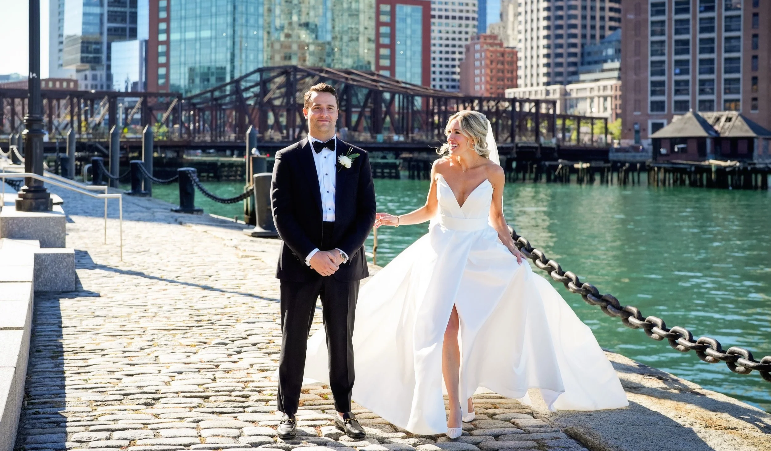 A newlywed couple in wedding attire standing on a cobblestone walkway by the water in Boston