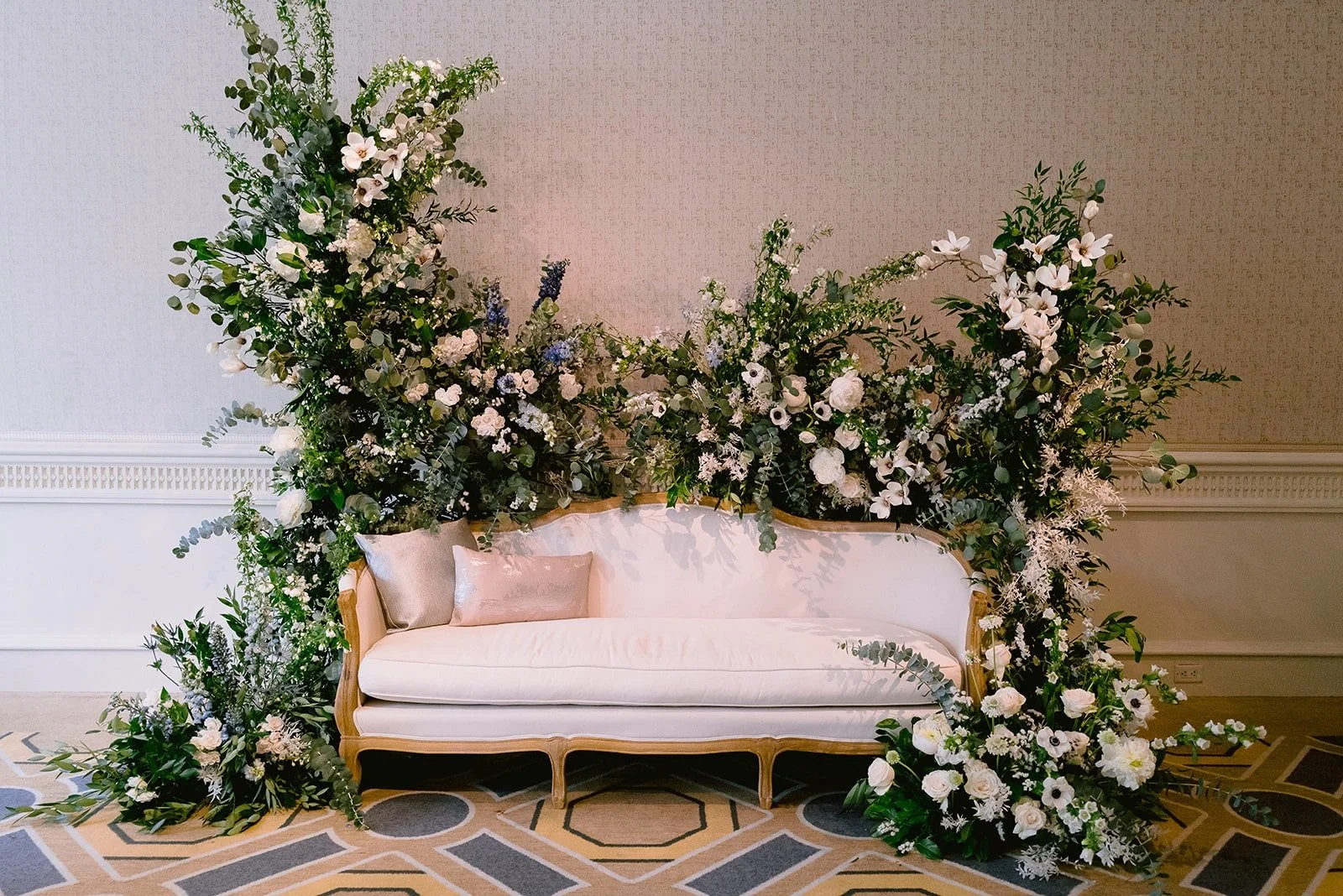 A white vintage sofa with gold frame and two decorative pillows, surrounded by an elaborate arrangement of white and green flowers and foliage, set against a plain wall in a decorated interior.