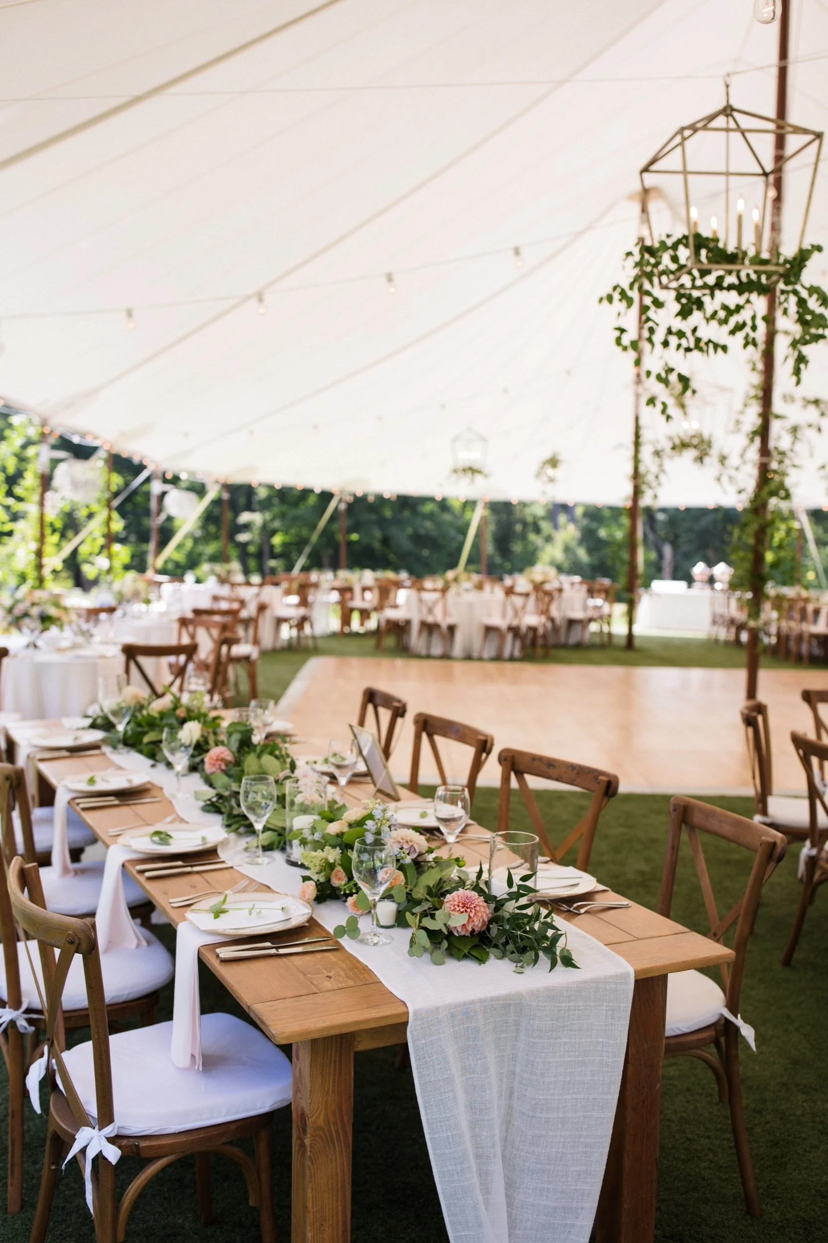Elegant outdoor event setup with long wooden tables decorated with white tablecloths, floral arrangements, glassware, and chairs, under a large white canopy with hanging string lights and greenery, in a lush garden setting.