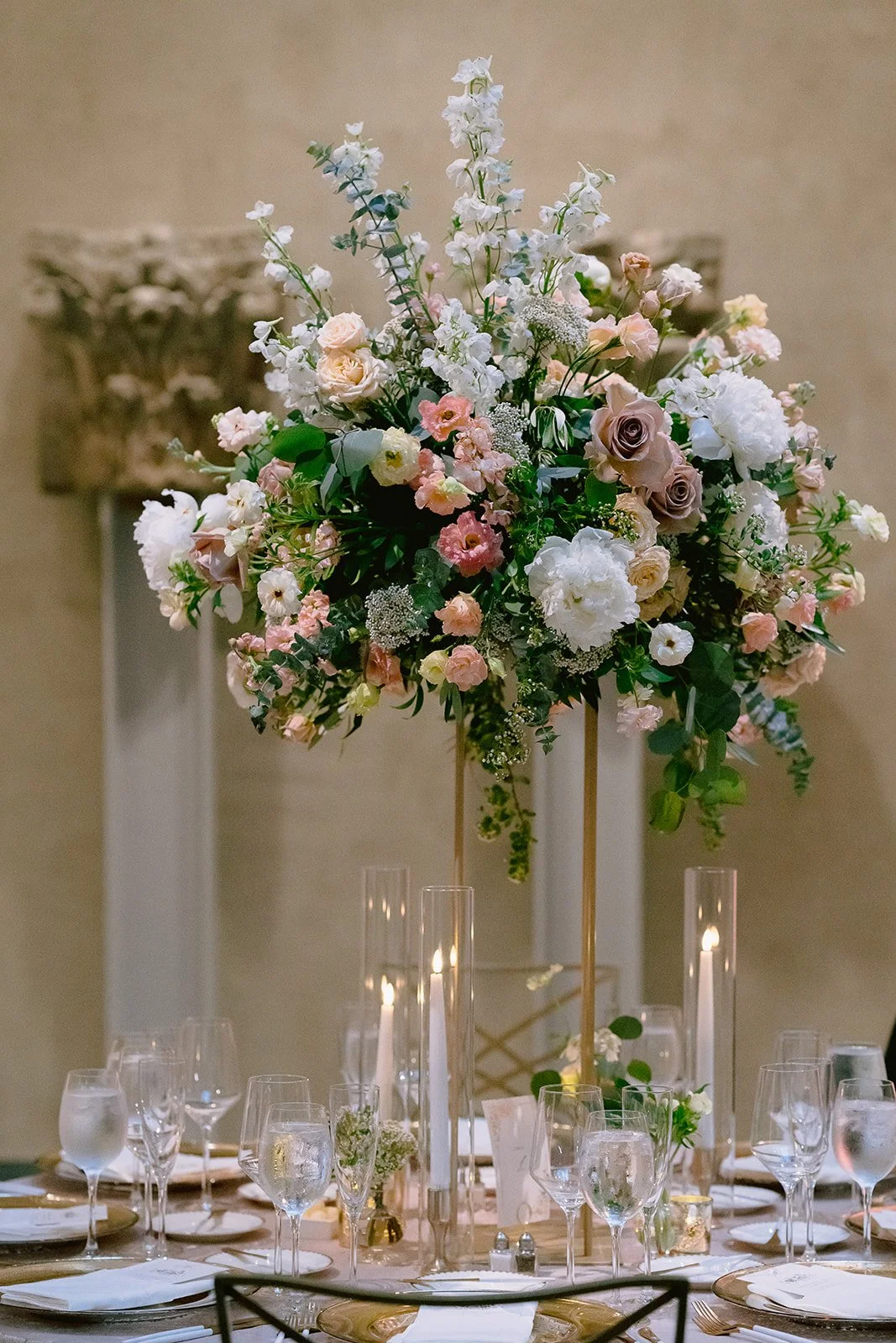 Elegant dining table with a large floral centerpiece featuring pink, white, and cream-colored flowers, along with green foliage, in tall gold stands. Surrounding the centerpiece are wine glasses, water glasses, candles, and place settings, indicating a formal event or celebration.