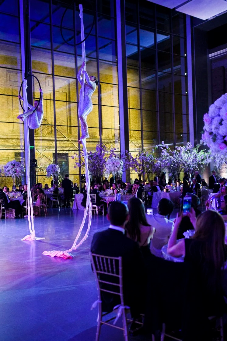 Two aerial silk performers in white costumes performing at an elegant event with guests seated at round tables decorated with floral centerpieces, inside a modern glass-walled venue illuminated with purple and yellow lighting.