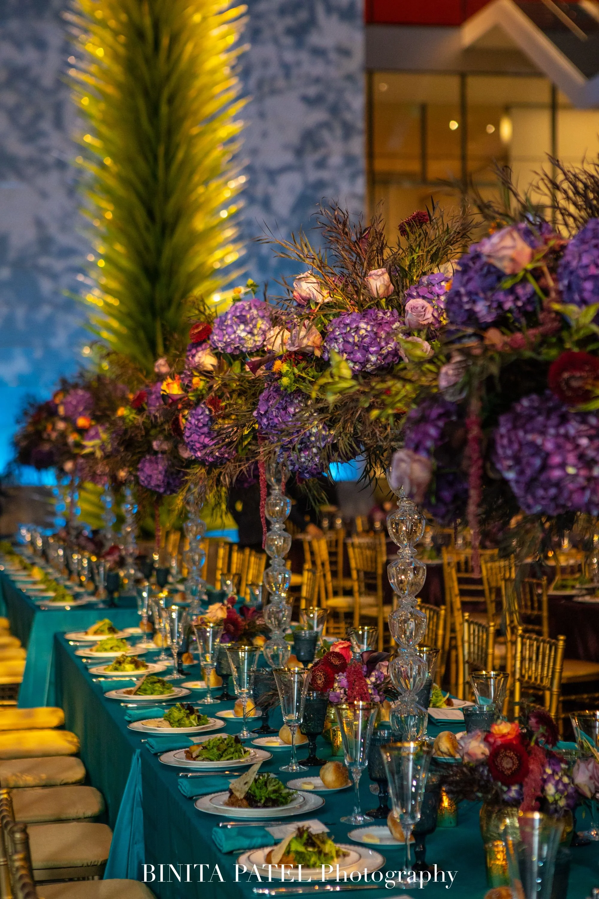 Elegant banquet table with tiered floral centerpieces, set with plates, glassware, and linens, in a decorated indoor setting.