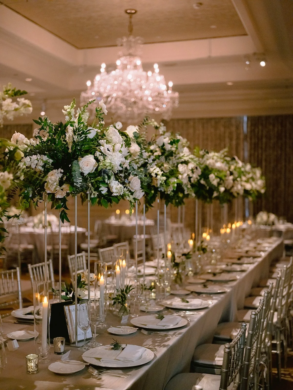 Wedding reception with a long banquet table decorated with white flowers and candles, a chandelier hanging from the ceiling, and elegant chairs.