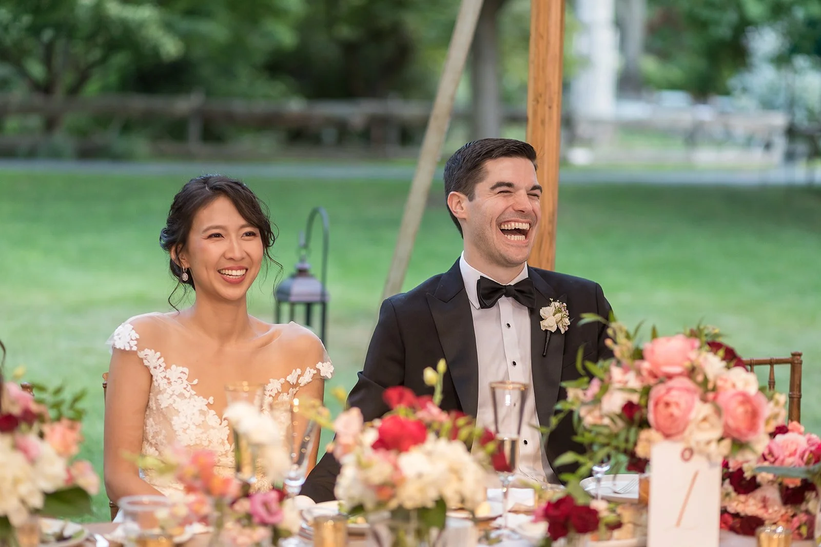 Happy bride and groom sitting at a wedding reception table outdoors, surrounded by flowers and greenery, smiling and laughing.