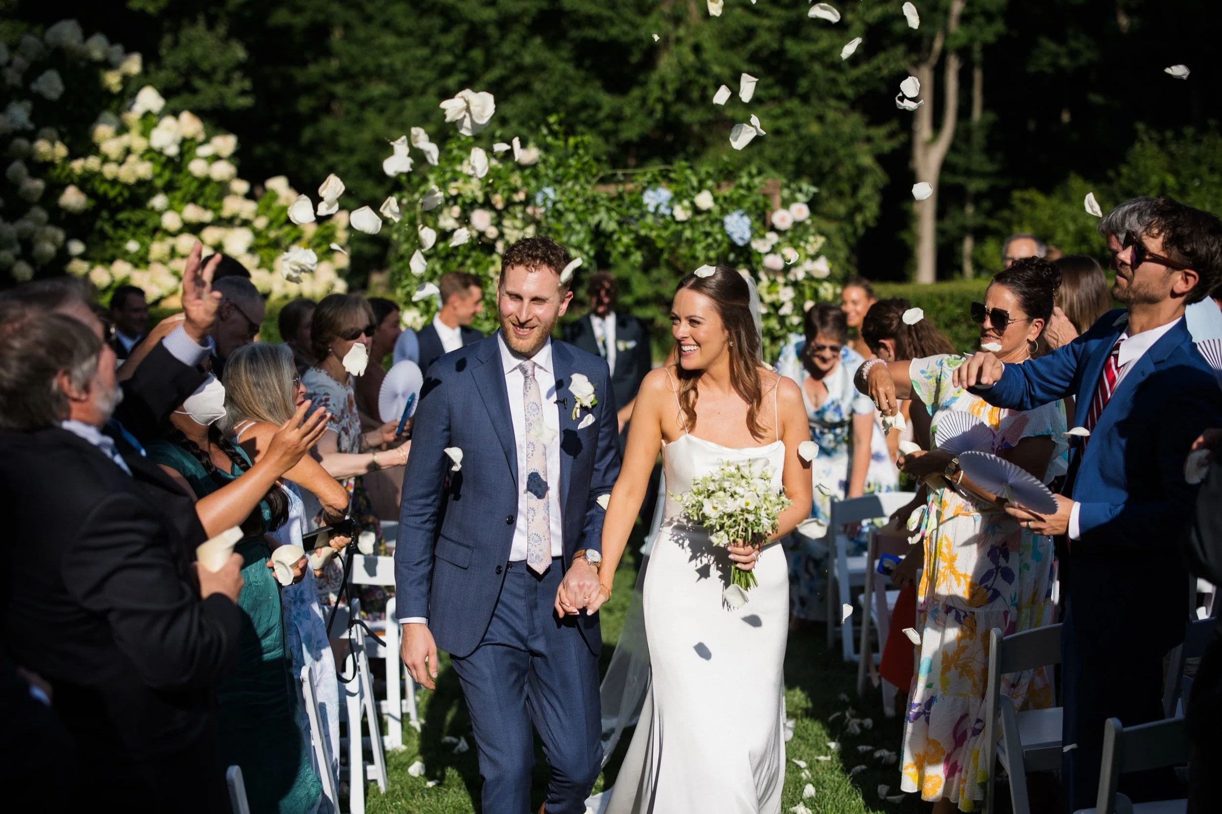 A bride and groom walking hand in hand down the aisle after their wedding ceremony outdoors, surrounded by smiling guests throwing flower petals.