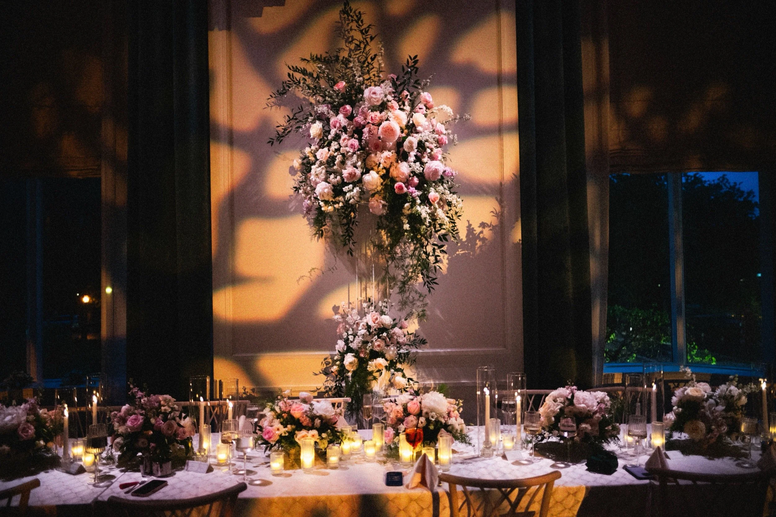 Elegant indoor wedding reception with a long table decorated with numerous pink and white flowers, candles, and wine glasses. A large floral arrangement is central behind the table, and blue evening sky can be seen through the windows.