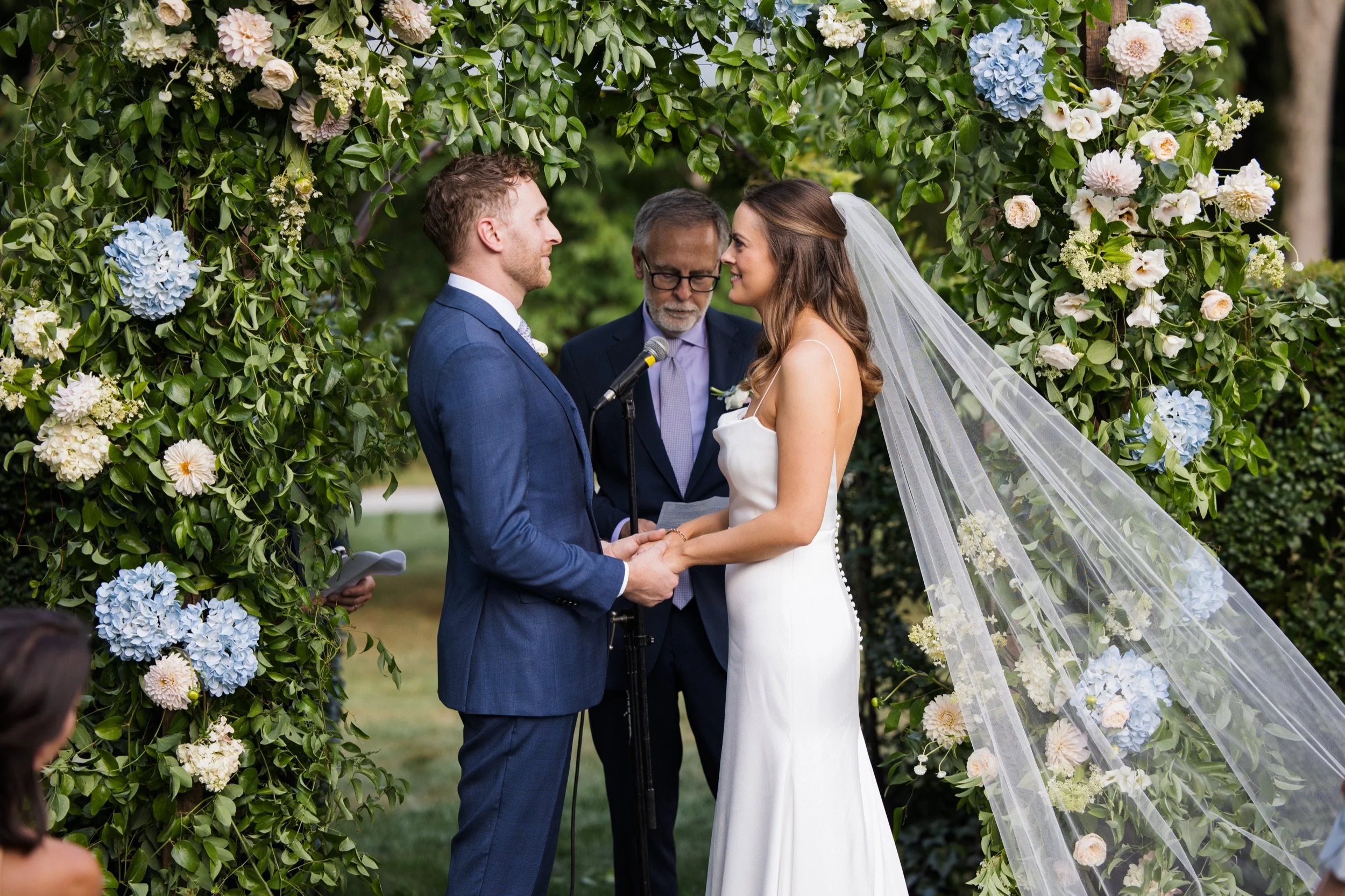 A bride and groom exchanging vows during an outdoor wedding ceremony under a floral arch with white and blue flowers, with an officiant standing between them.