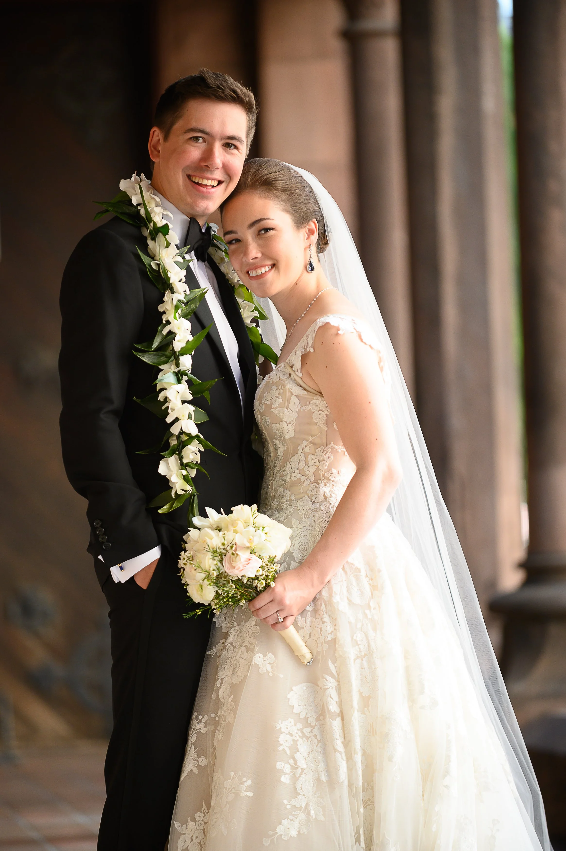 A smiling bride and groom standing close together in wedding attire, with the bride holding a bridal bouquet, inside a building with tall columns in the background.