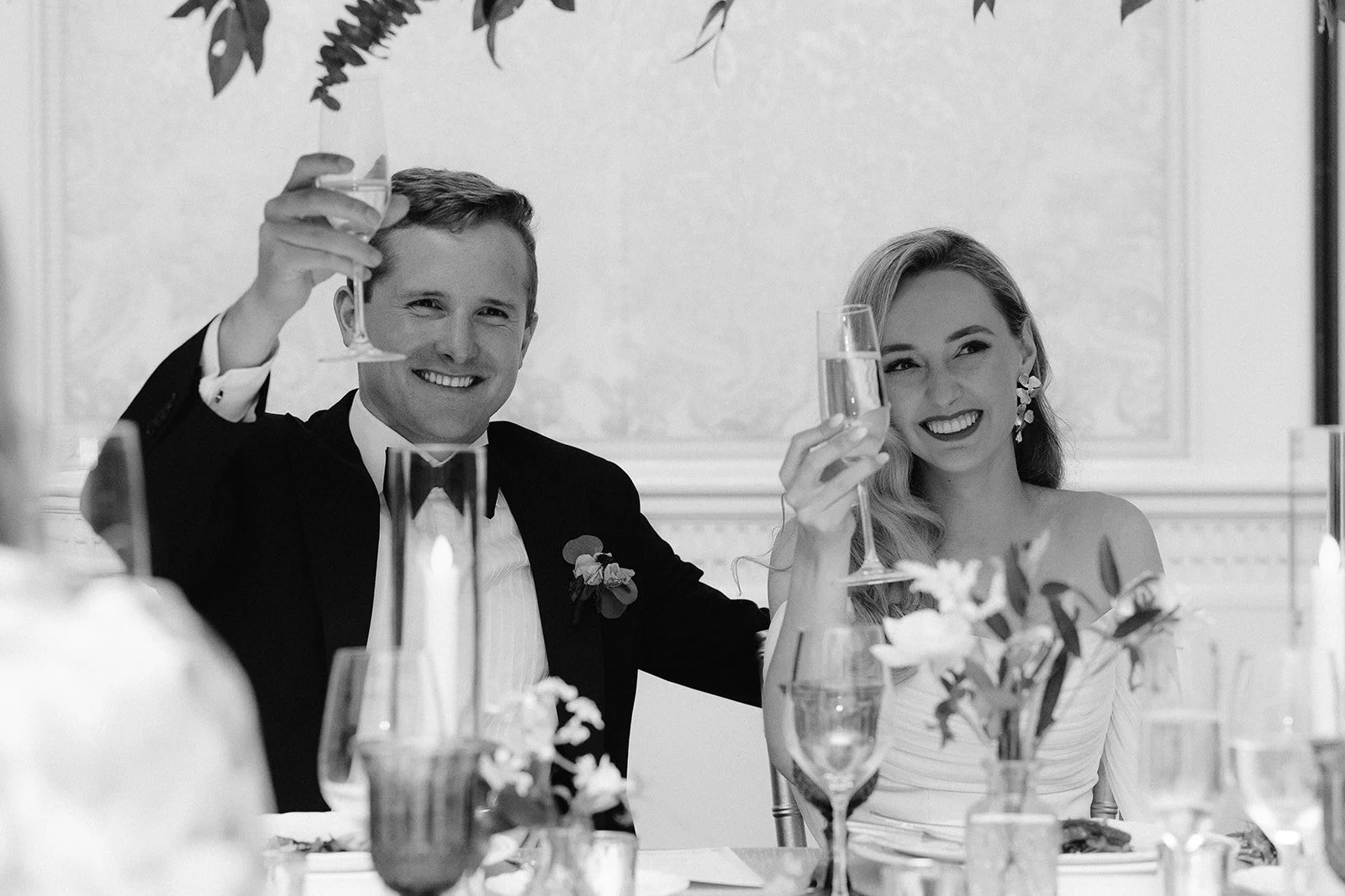 Black and white photo of a happy couple at a wedding reception, raising glasses of champagne for a toast, sitting at a table with flowers and candles.