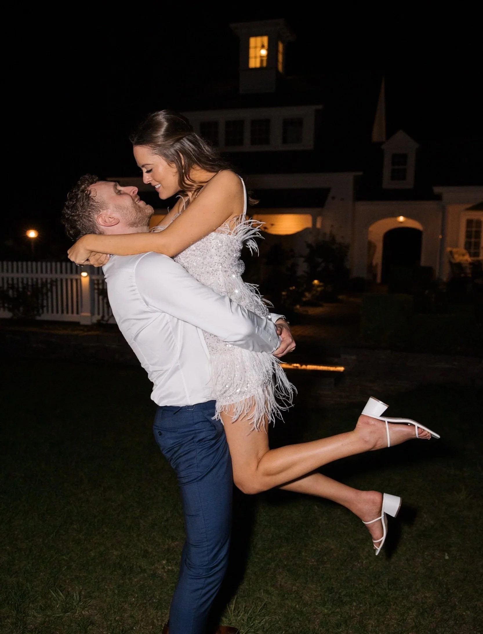A man is holding a woman in the air at night outside. She is smiling, leaning toward him with her legs wrapped around his waist and her arms around his neck. She is wearing a white dress and high heels. The man is wearing a white shirt and dark pants.