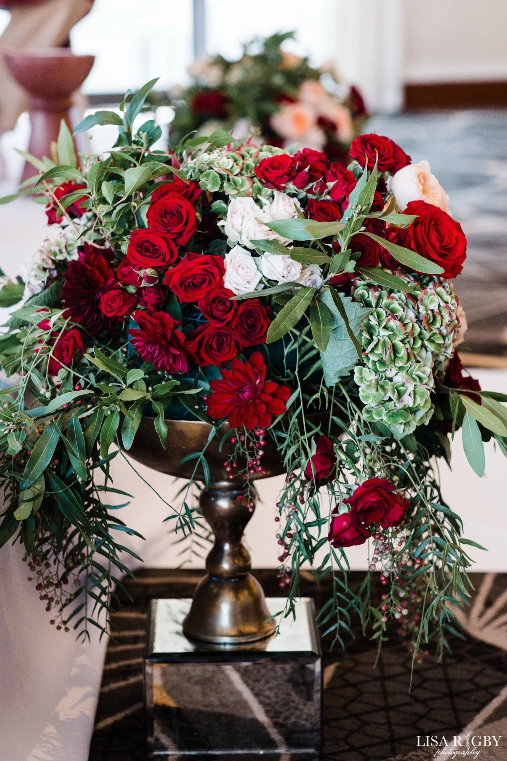 A floral arrangement in a gold vase sitting on a black table, with red and white roses, green leaves, and pinkish flowers, in a room with patterned carpet and a white wall in the background.