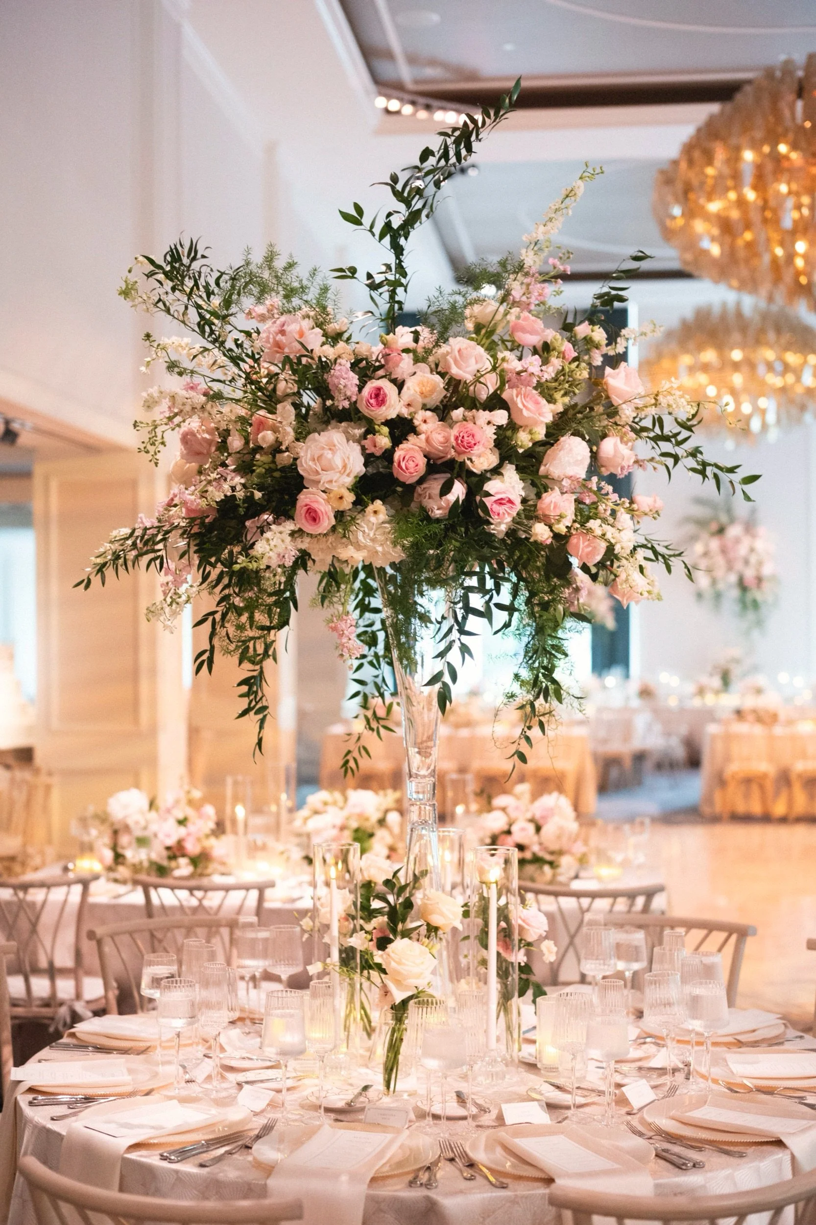A tall floral arrangement of pink and white roses, greenery, and small white flowers on a wedding reception table, with candles and table settings in the background.