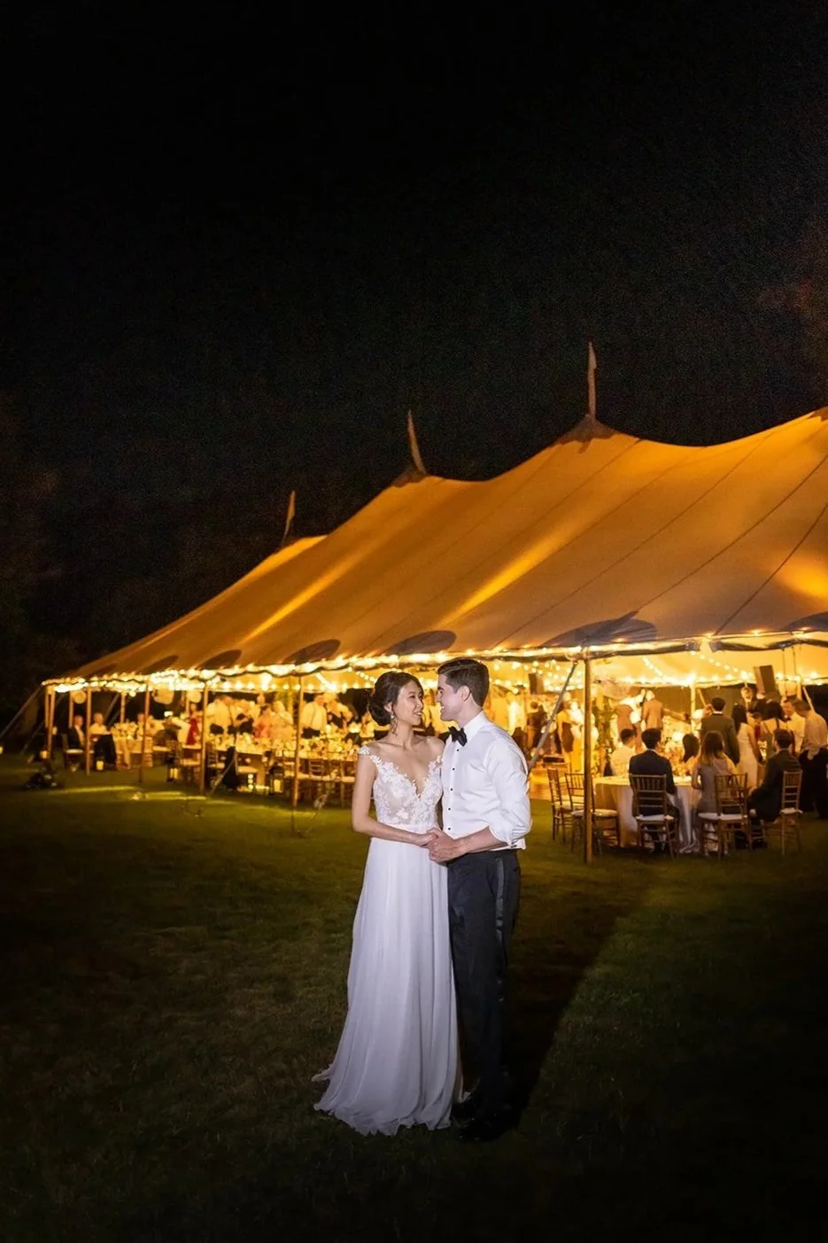 A bride and groom holding hands at night outdoors during their wedding reception, with a lit tent and guests seated at tables in the background.