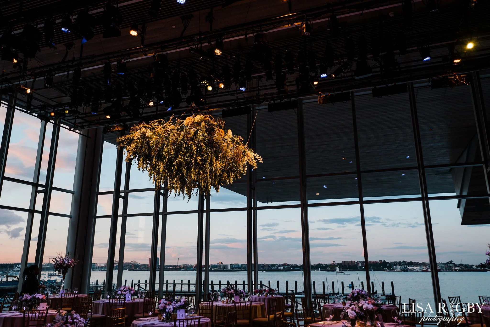 Indoor event space with large windows overlooking a waterfront, decorated with floral arrangements and set dining tables, under a ceiling with stage lighting and a hanging flower decoration.