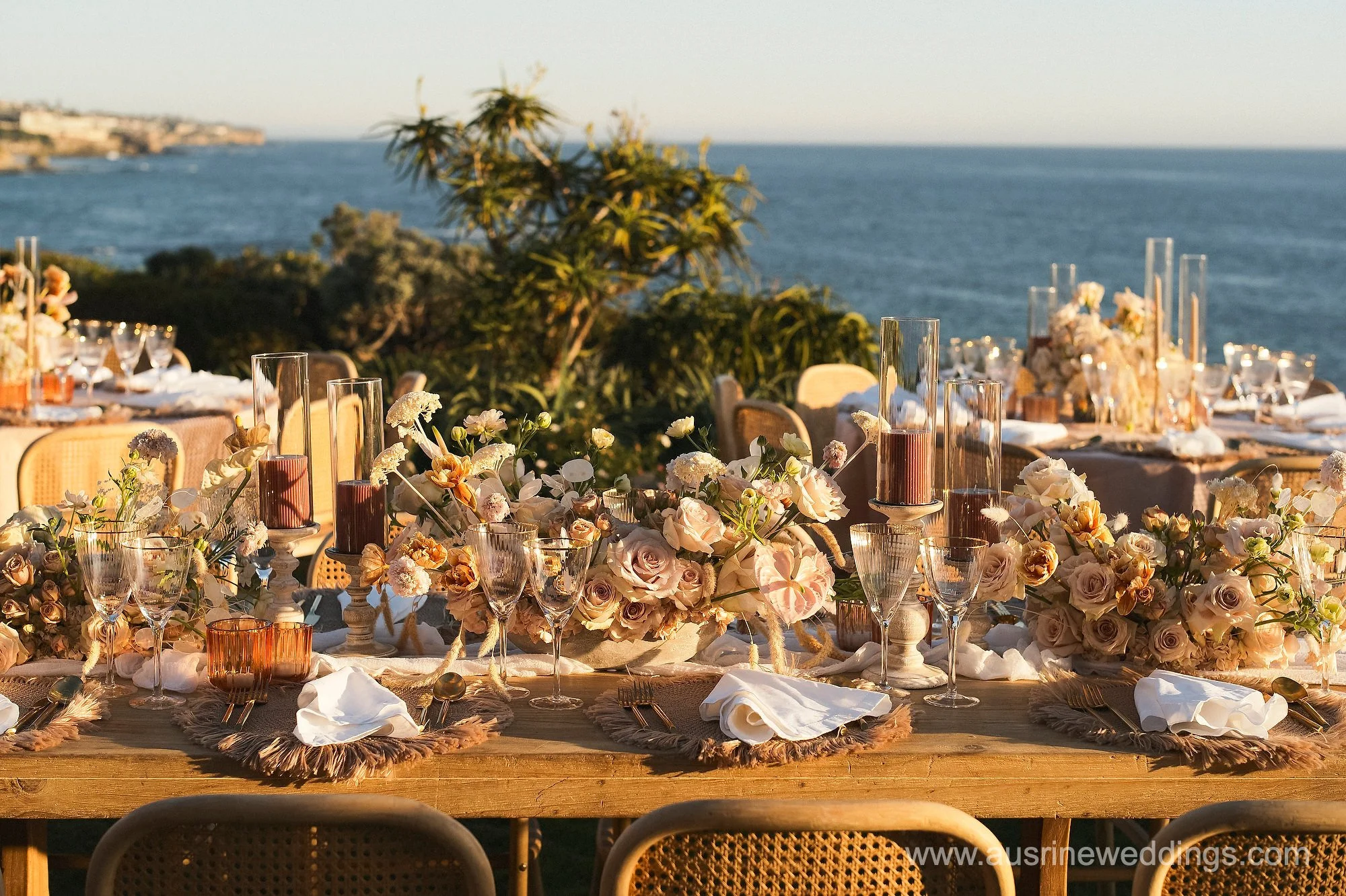 A long outdoor dining table decorated with flowers, candles, and glassware, overlooking the ocean at sunset.