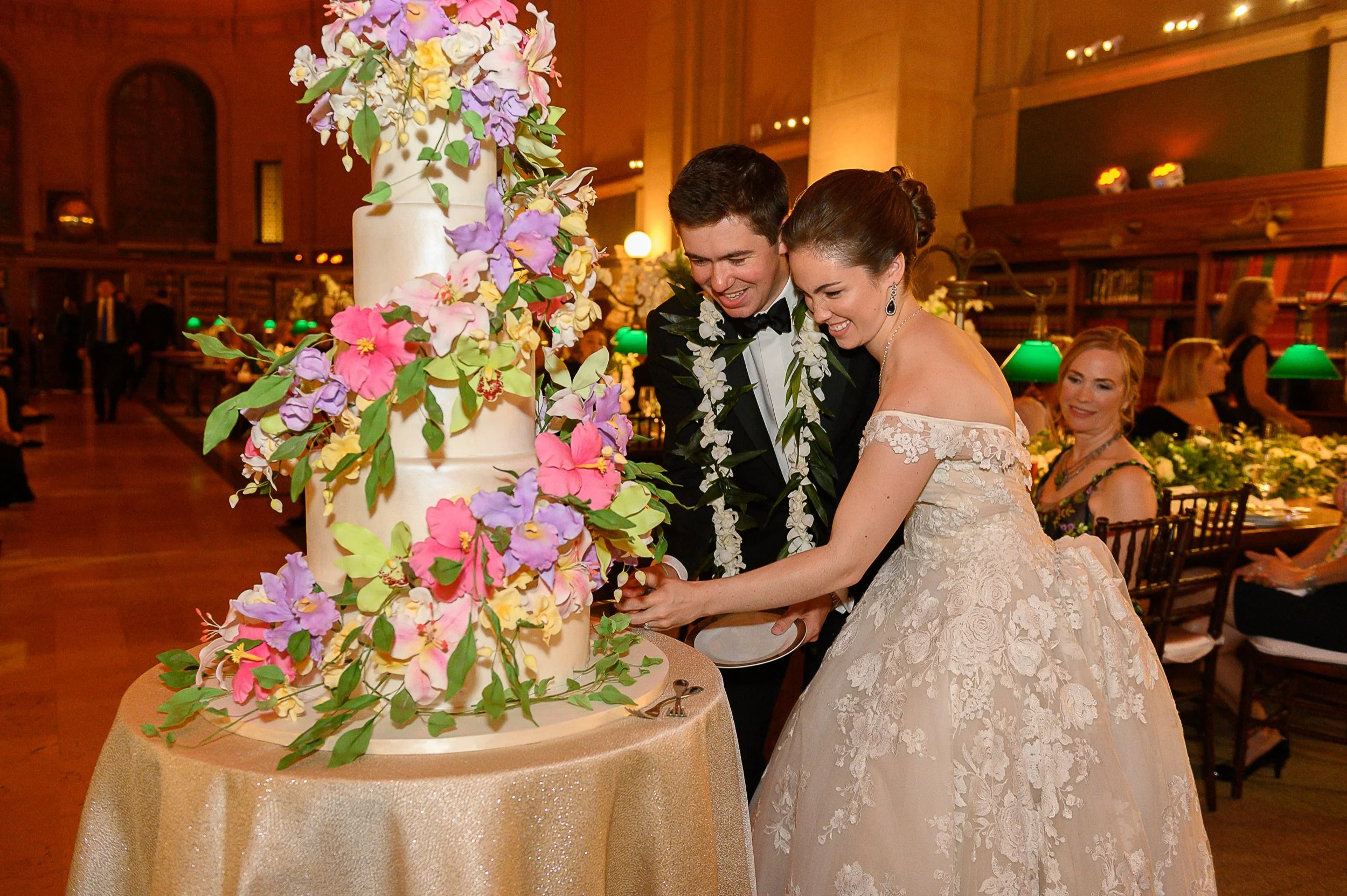 A bride and groom cutting a wedding cake together at a wedding reception. The cake is decorated with colorful flowers and the couple is smiling and wearing wedding attire. The background shows guests and elegant decor.