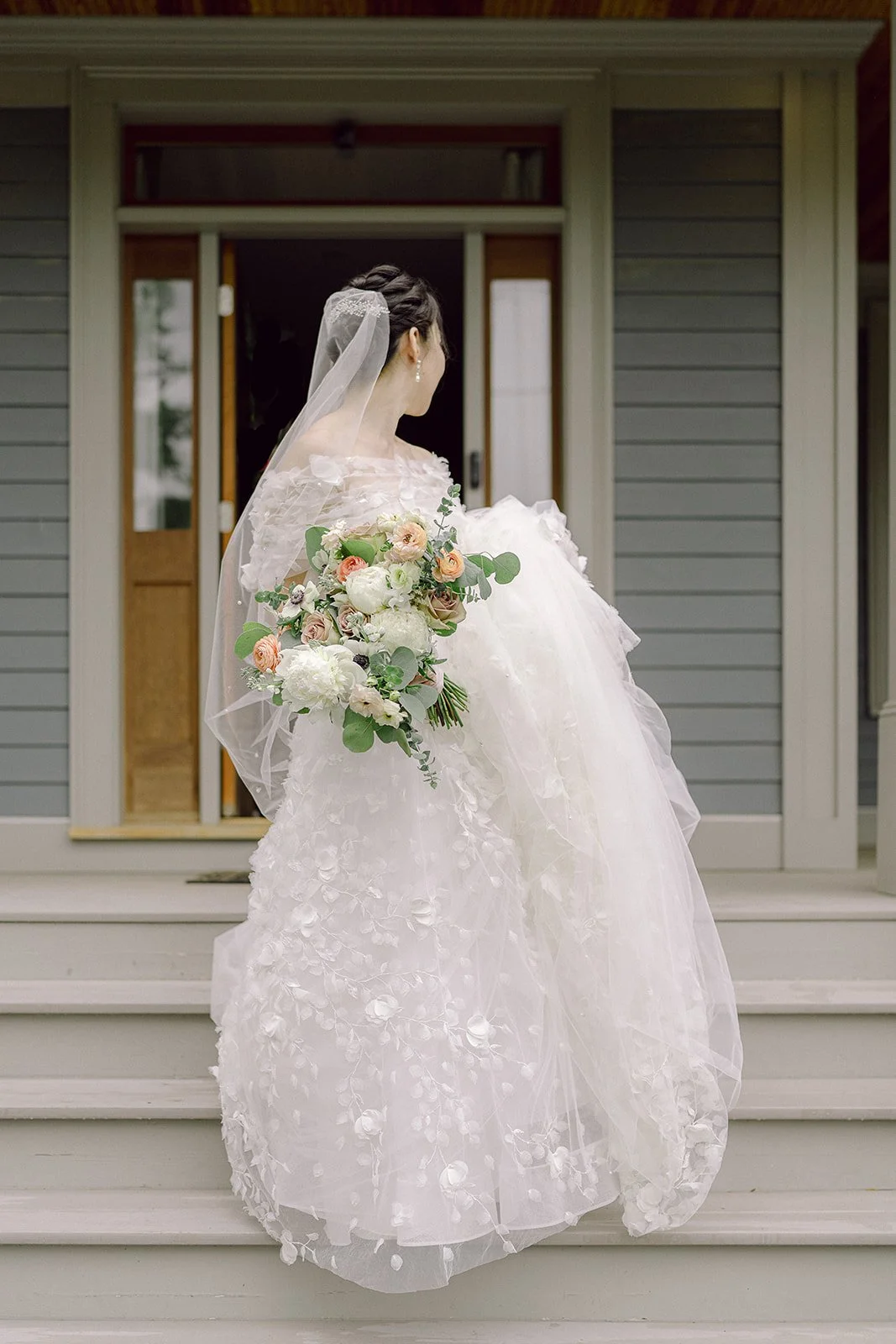 Bride in a white wedding gown holding a large bouquet of flowers, standing on the steps outside her house, facing away from the camera.