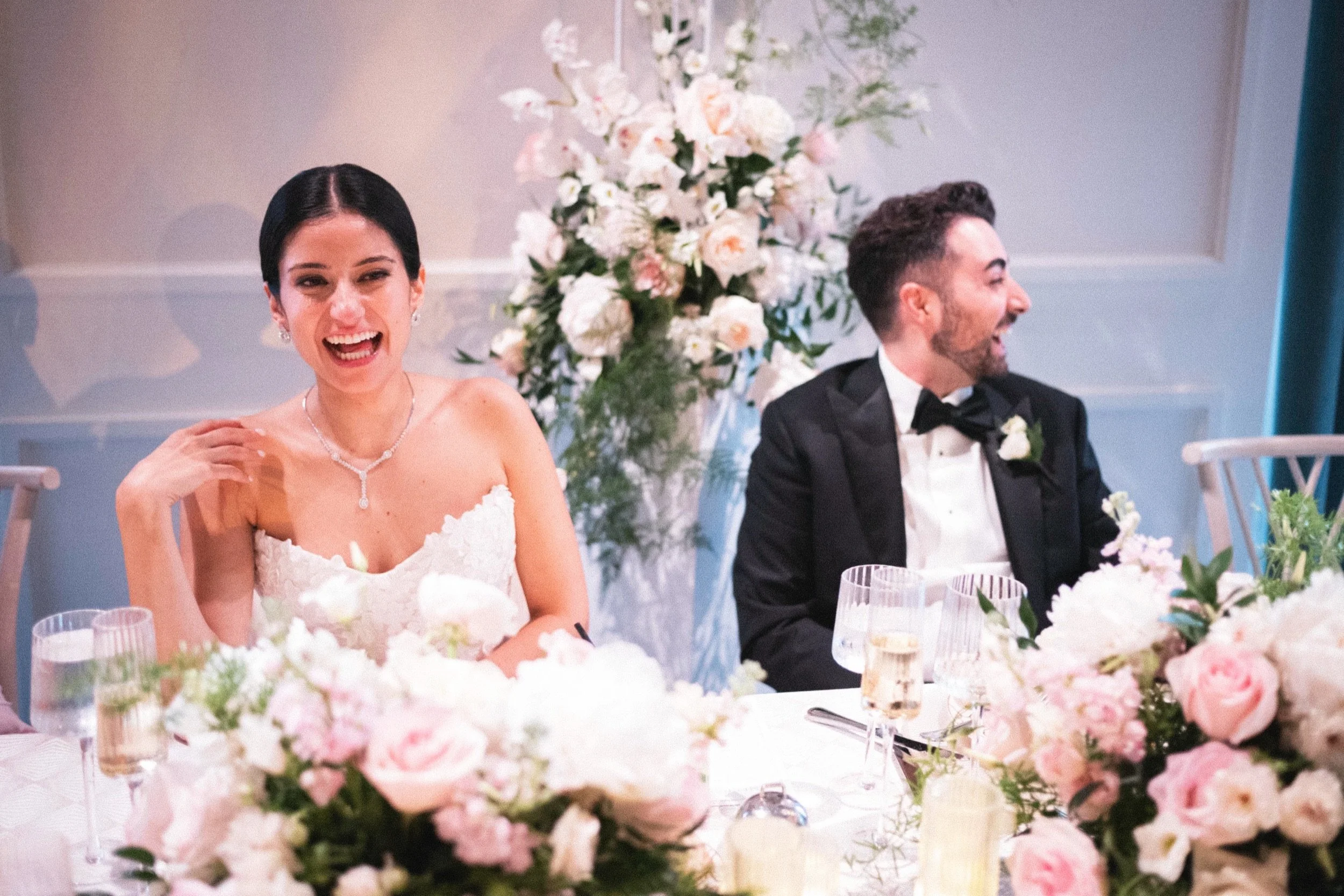 A bride and groom sitting at a wedding reception table decorated with pink and white flowers, smiling and enjoying the moment.