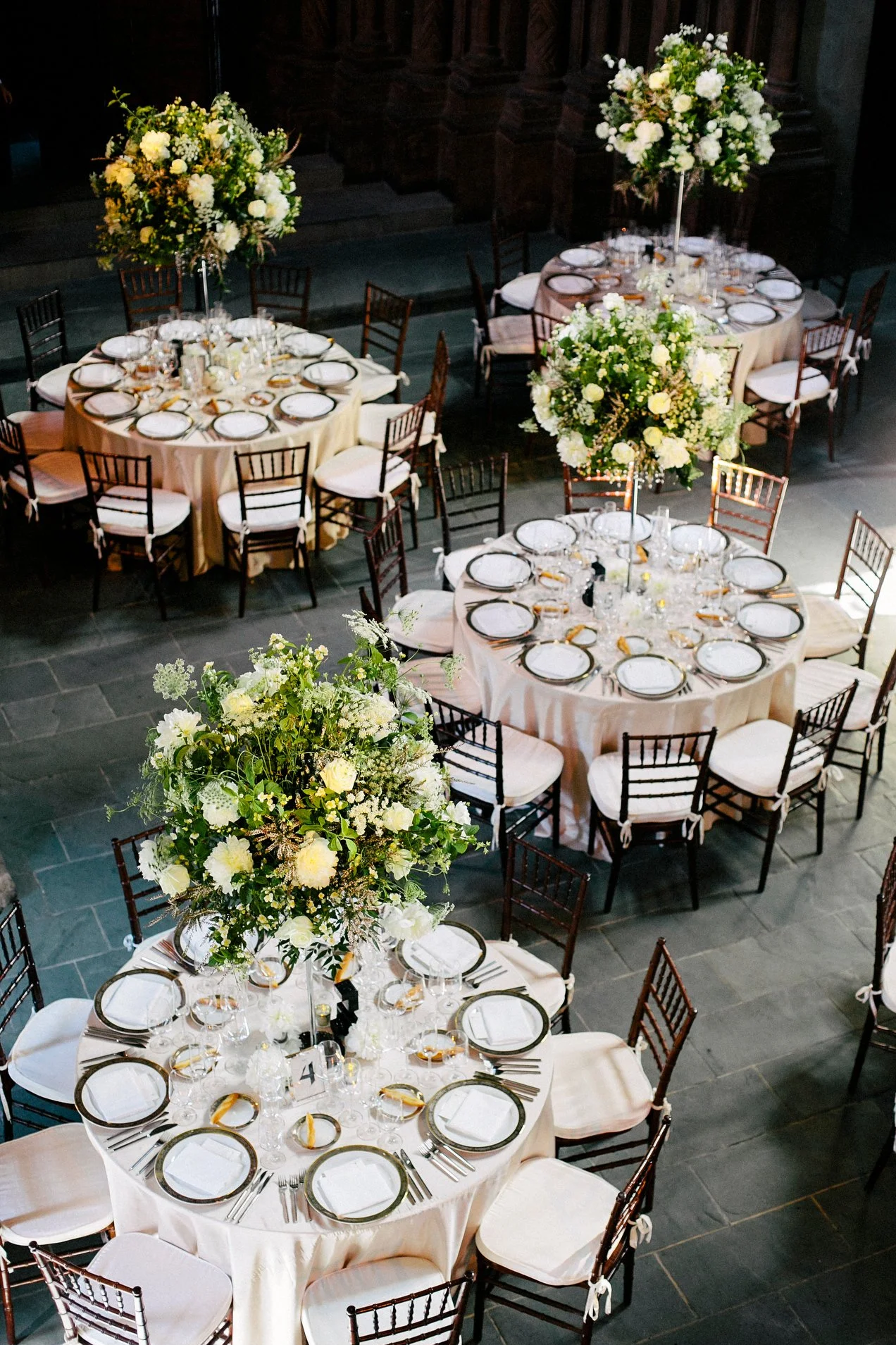 Elegant banquet hall with round tables covered in cream tablecloths, set with white and gold-rimmed dinnerware, and tall floral centerpieces of white flowers on tall stands.
