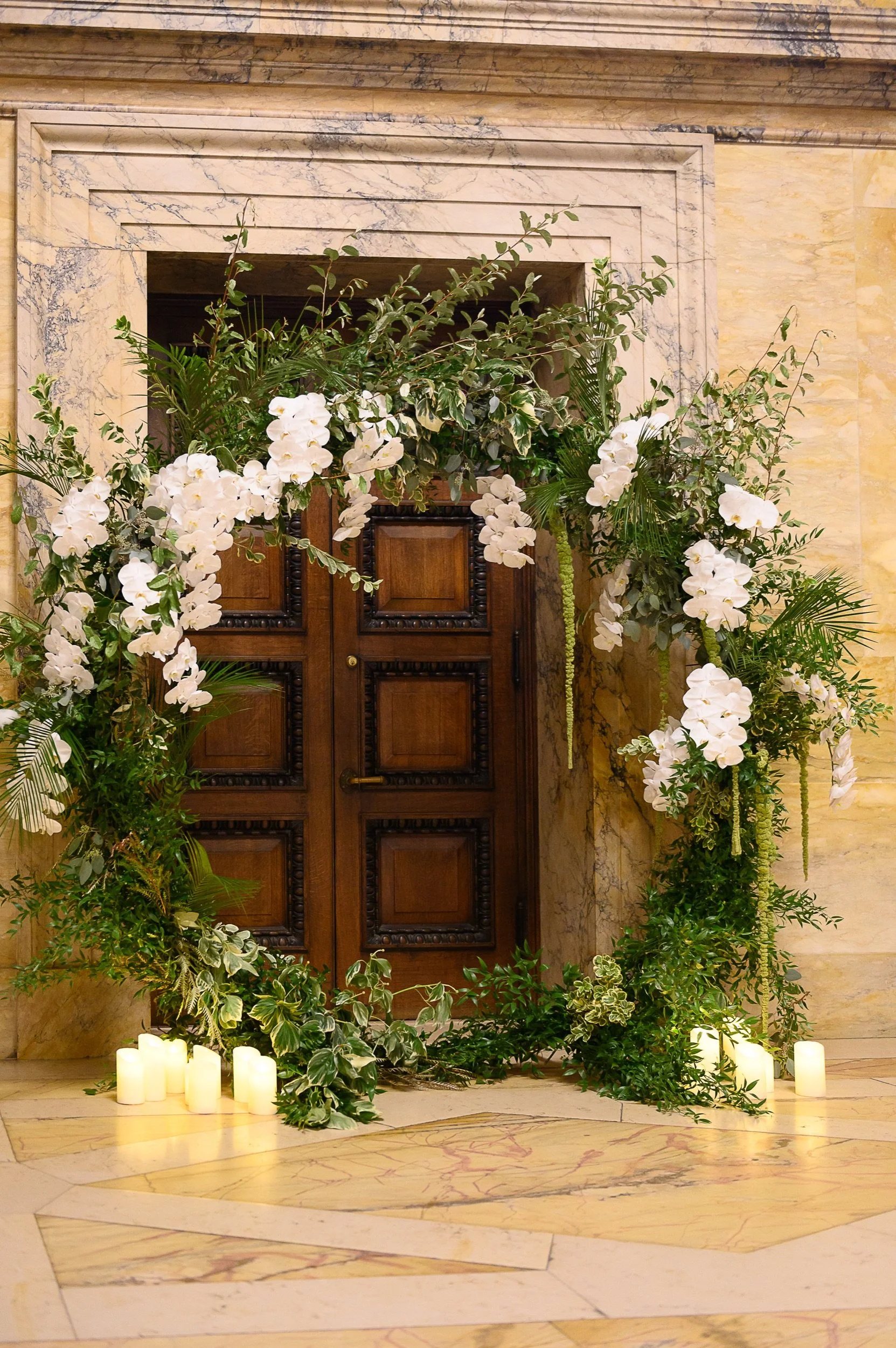 Floral arrangement with white orchids, green leaves, and candles in front of a wooden door, set against marble walls and floor.