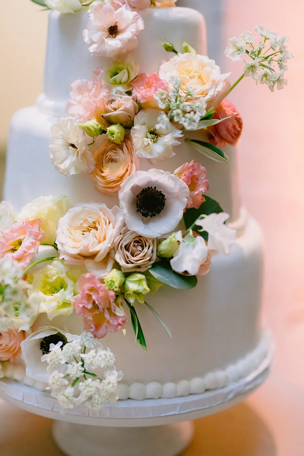 A white tiered cake decorated with various pastel-colored flowers including roses, anemones, and lisianthus.