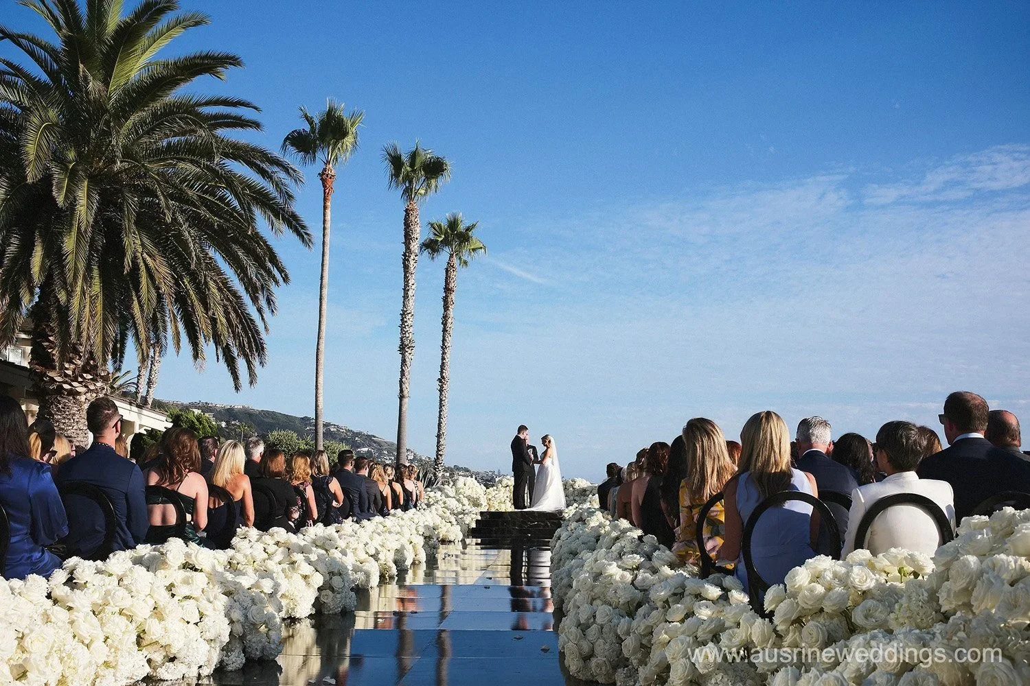 Outdoor wedding ceremony on a sunny day featuring a bride and groom standing together under the sky, surrounded by guests seated along the aisle decorated with white flowers, with palm trees in the background.