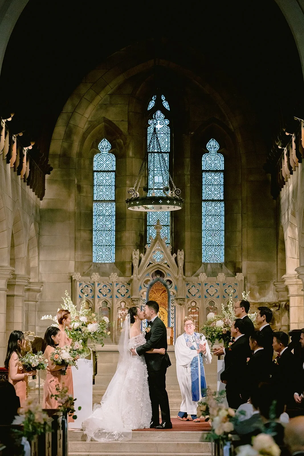A wedding ceremony taking place inside a church with a bride and groom kissing at the altar, surrounded by bridesmaids, groomsmen, and an officiant, with stained glass windows and floral arrangements in the background.