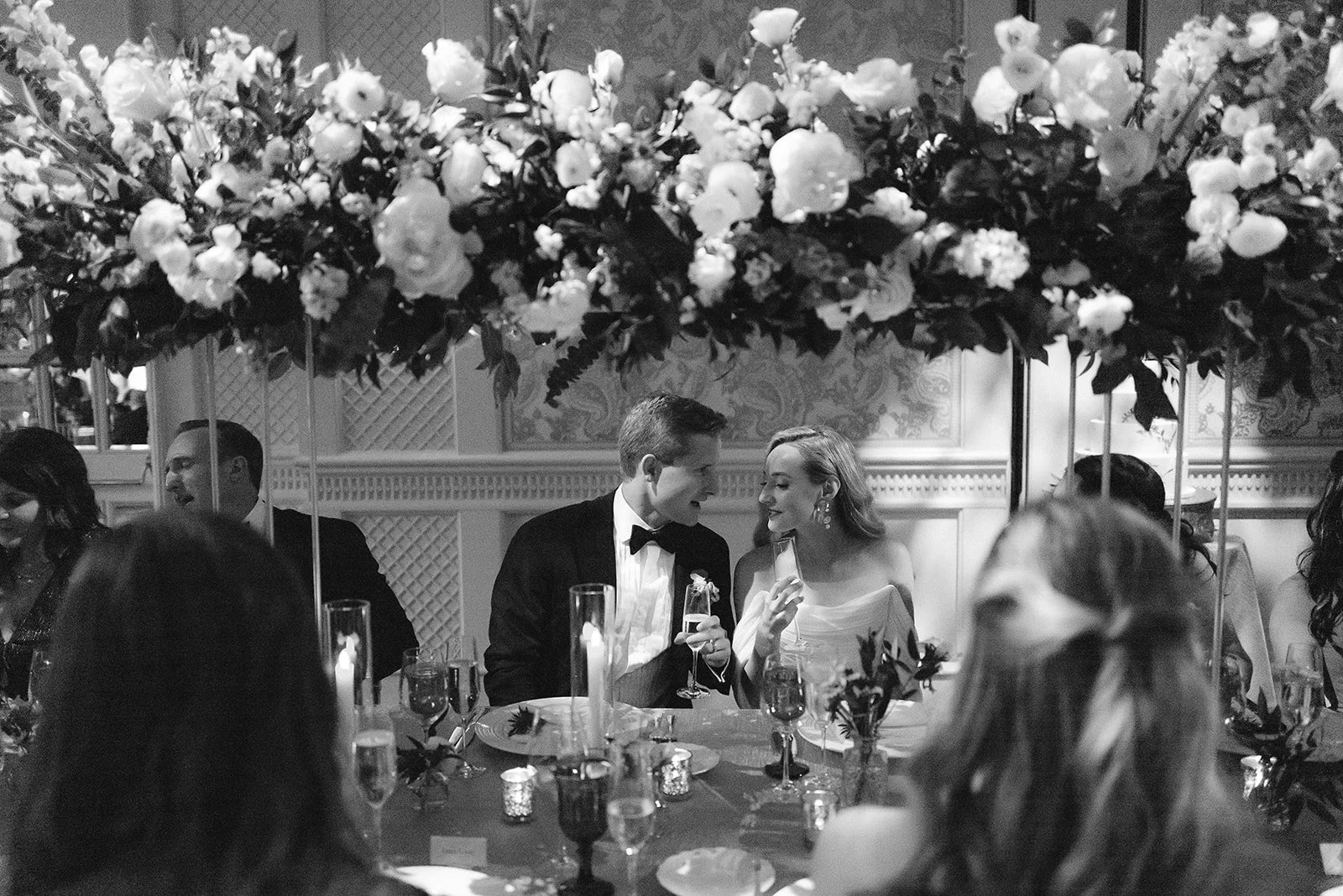 A black-and-white photo of a formal wedding reception with a long table decorated with flowers and candles. A couple in wedding attire, a groom in a tuxedo and a bride in a white dress, are seated together, smiling and sharing a drink. Other guests are visible around the table, and a large floral arrangement hangs above them.