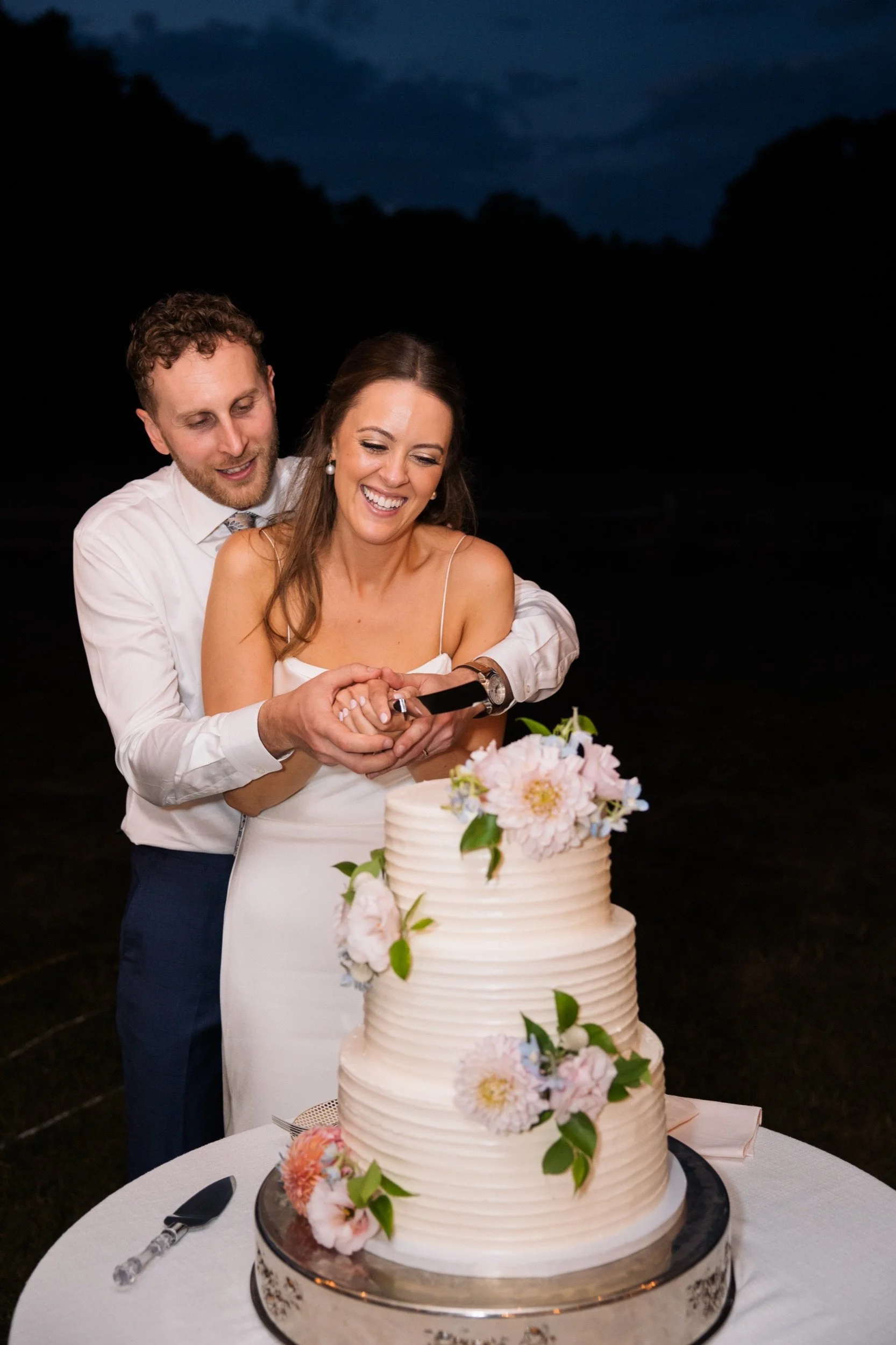 A happy couple cutting a wedding cake outdoors at night. The cake is decorated with pink and white flowers and green leaves, placed on a silver stand on a white table with a cake serving knife and fork.