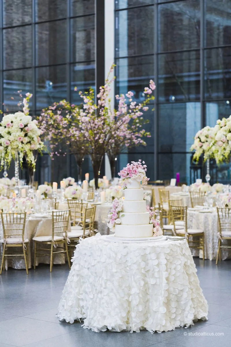 A wedding reception event with decorated tables, flowers, and a wedding cake on a table in the foreground, inside a modern building with large glass windows.