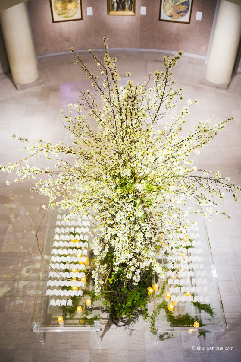 A large floral arrangement with white blossoming branches in a clear glass container, surrounded by small yellow candles and greenery, displayed on a glass table in an art gallery setting.