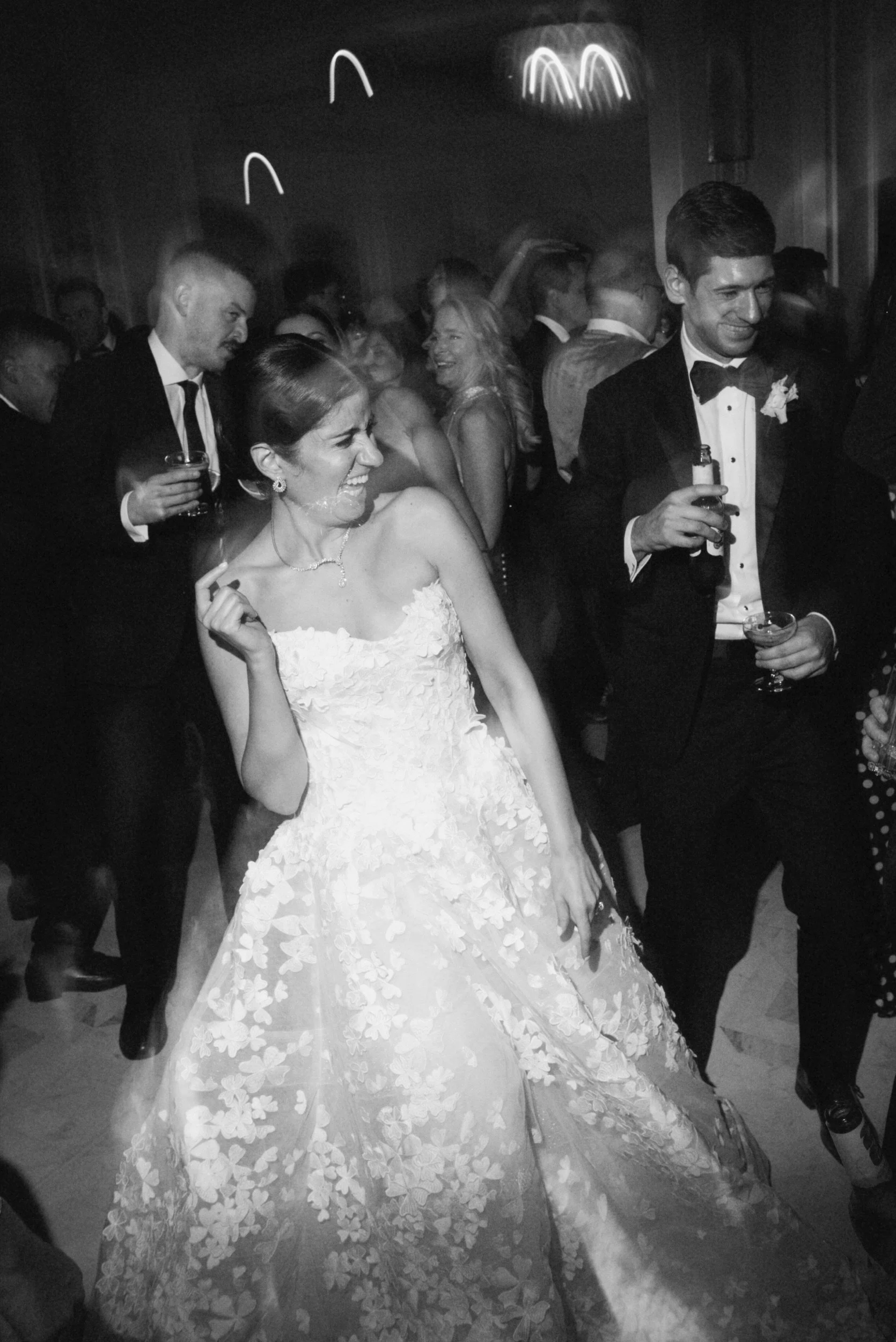 People dancing and celebrating at a wedding reception, with a bride in a white gown and guests in tuxedos and formal dresses in a dimly lit room.