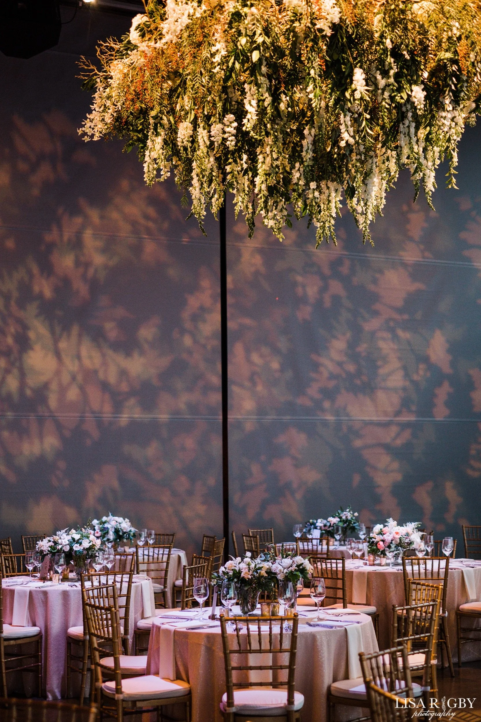 Elegant banquet hall decorated with floral centerpieces, round tables with white tablecloths, and gold Chiavari chairs, illuminated by warm lighting. A floral chandelier hangs from the ceiling, casting shadows on a dark wall.