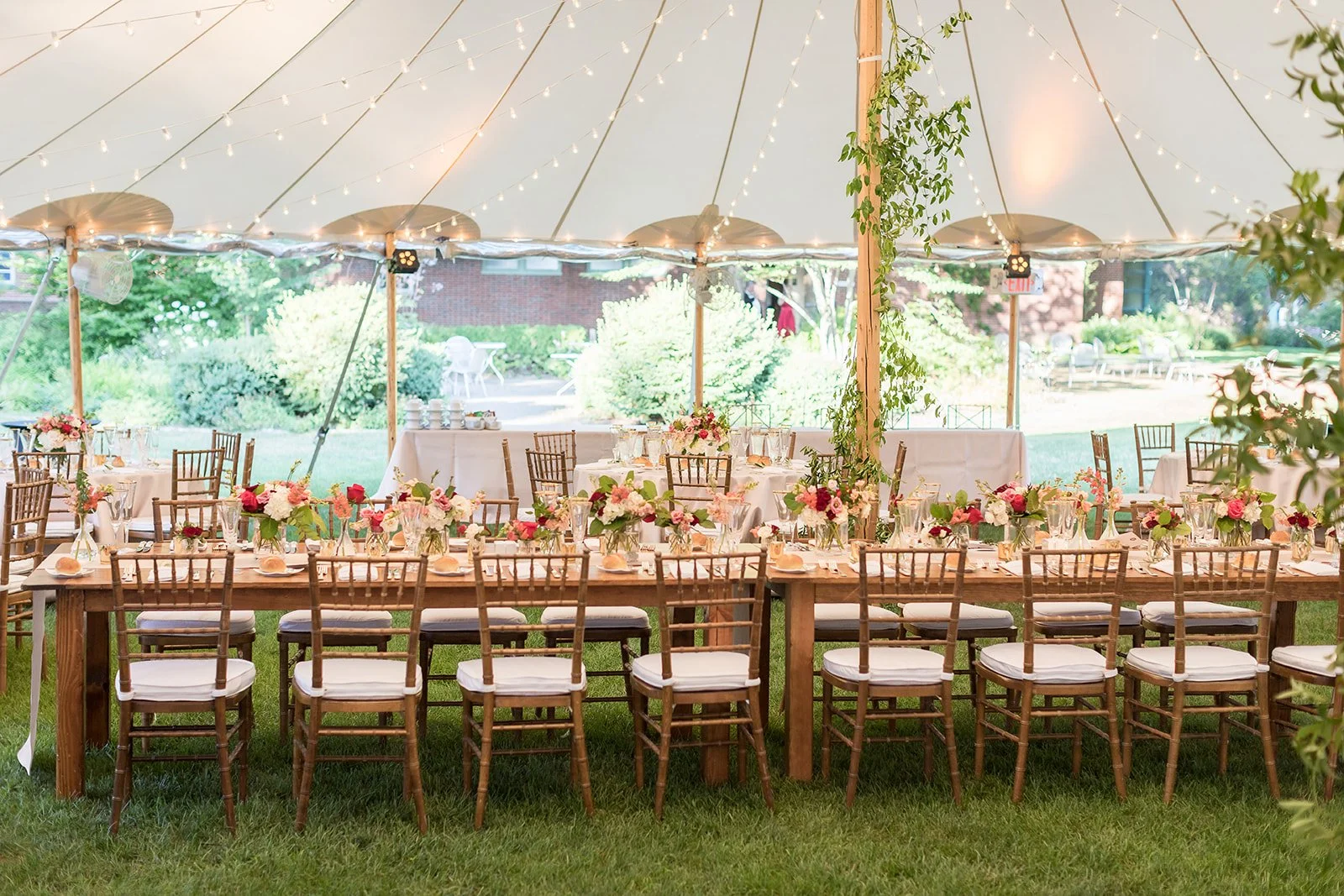 A decorated outdoor event tent with a long wooden table, adorned with floral centerpieces in glass vases, set with plates and utensils, surrounded by wooden chairs with white cushions. String lights hang from the tent ceiling, and there are lush green bushes and trees in the background.