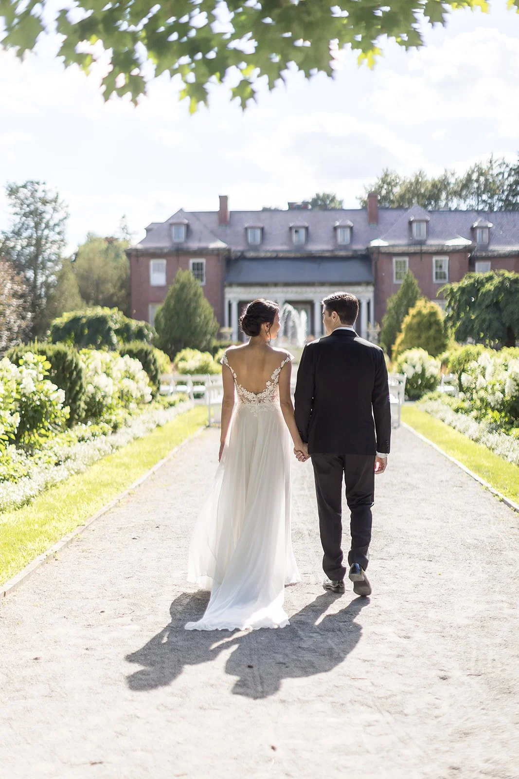 A bride and groom holding hands walk along a garden pathway toward a large mansion, surrounded by lush greenery and blooming flowers on a sunny day.