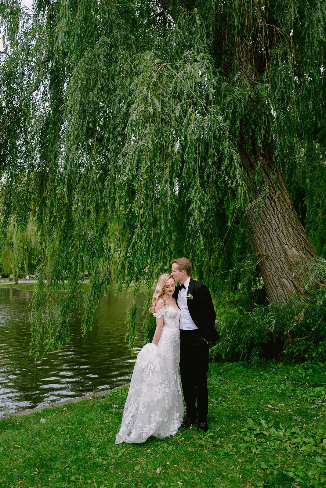 A bride and groom standing by a lake with a large weeping willow tree in the background.
